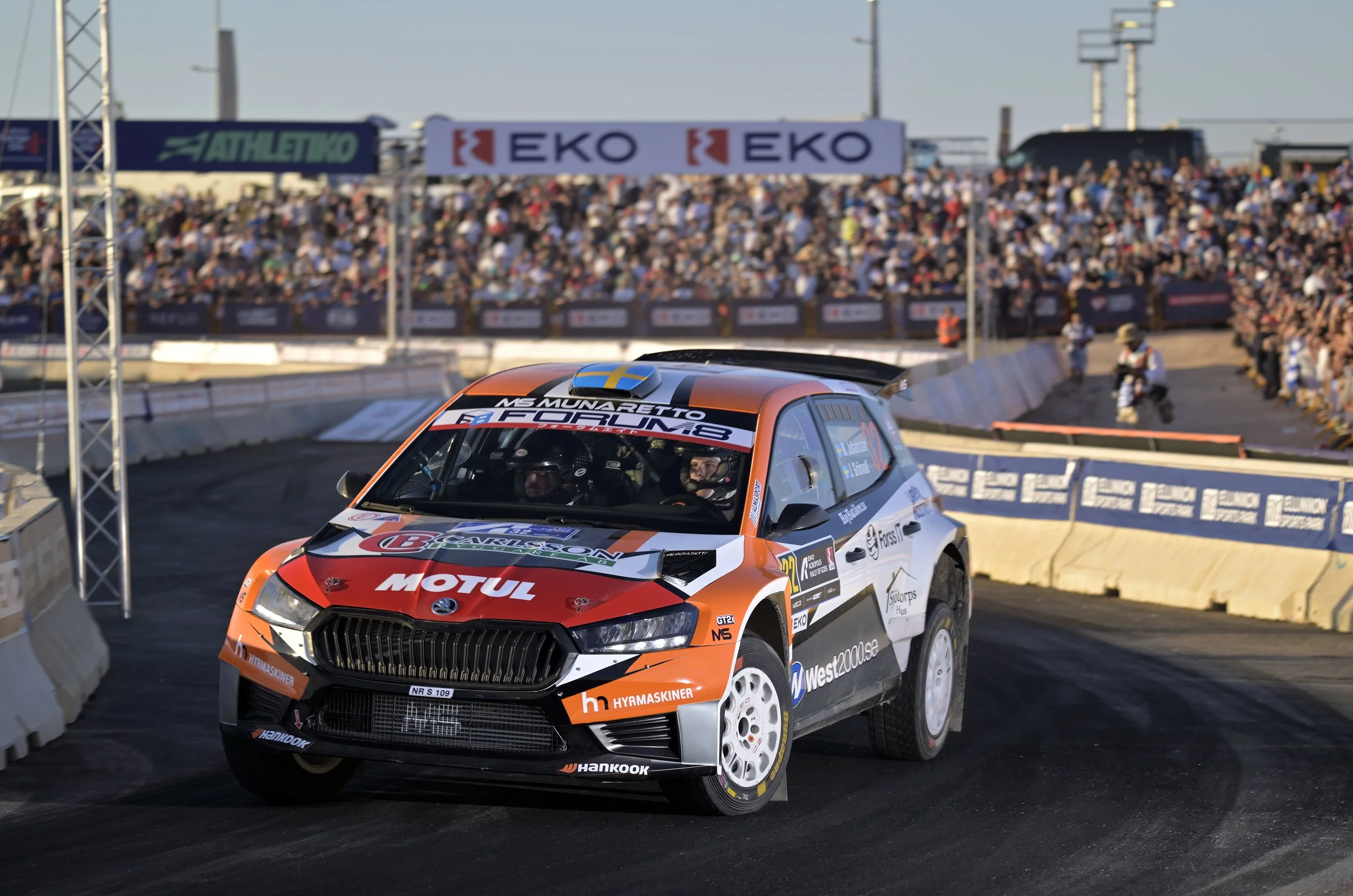 A rally car with orange, white, and black livery on a race track, approaching a corner as a large crowd watches from the stands in the background.
