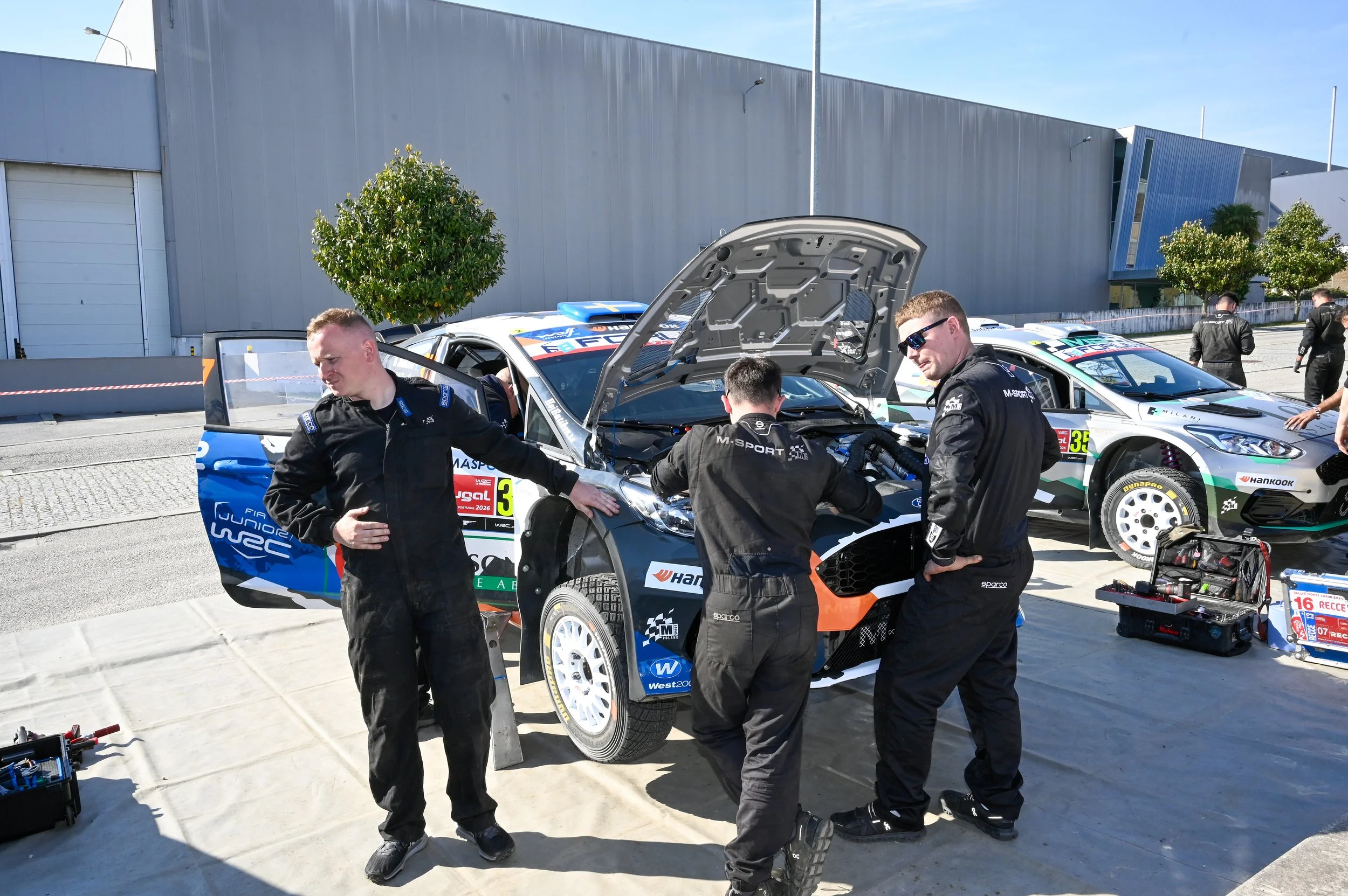 Race car team members working on a rally car in a pit area, with some standing around and others inspecting the engine with the hood open. The scene takes place outdoors on a sunny day with a large industrial building and trees in the background.