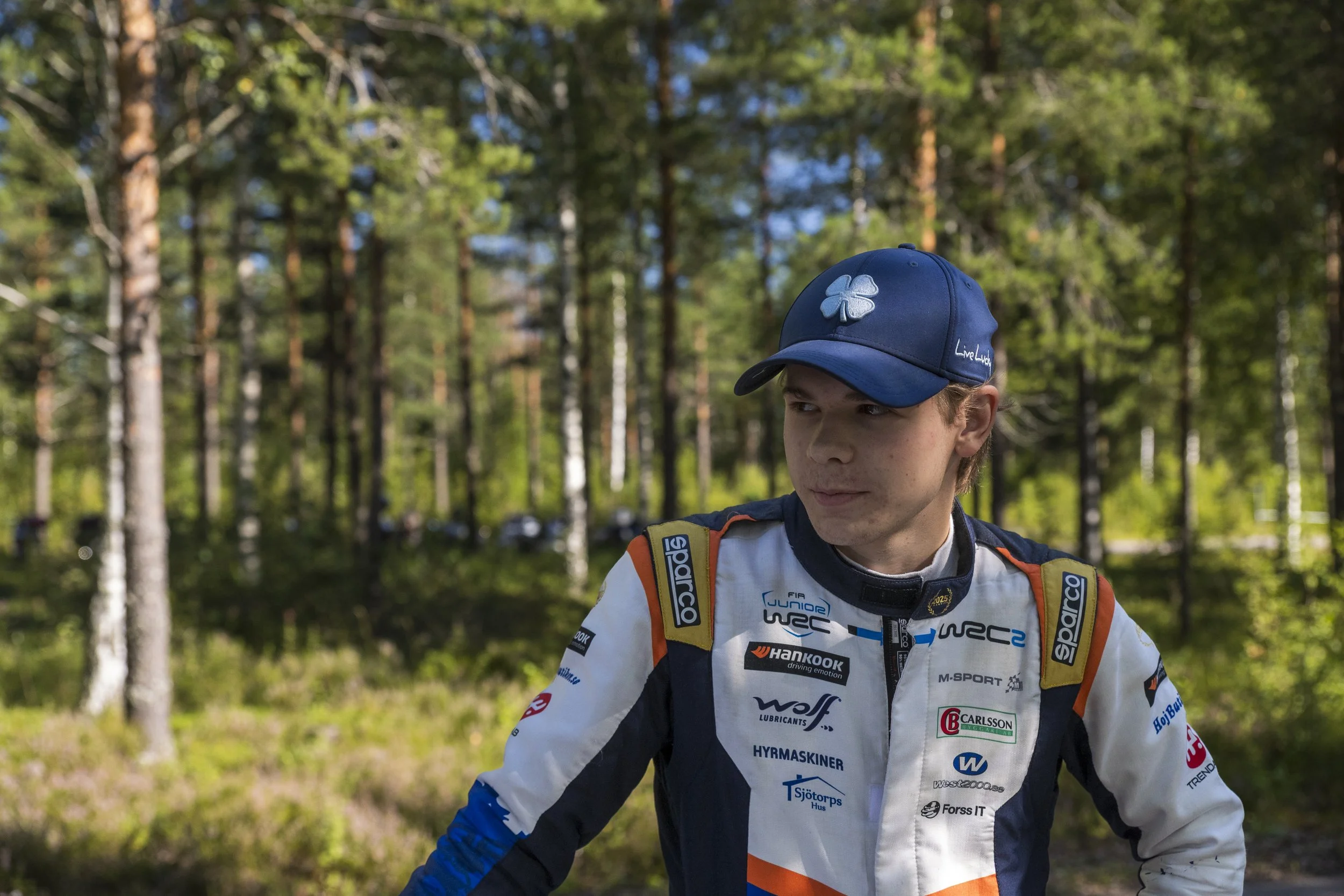 A young race car driver in a white racing suit with sponsor logos, wearing a blue cap with a four-leaf clover emblem, standing outdoors among trees in a wooded area.