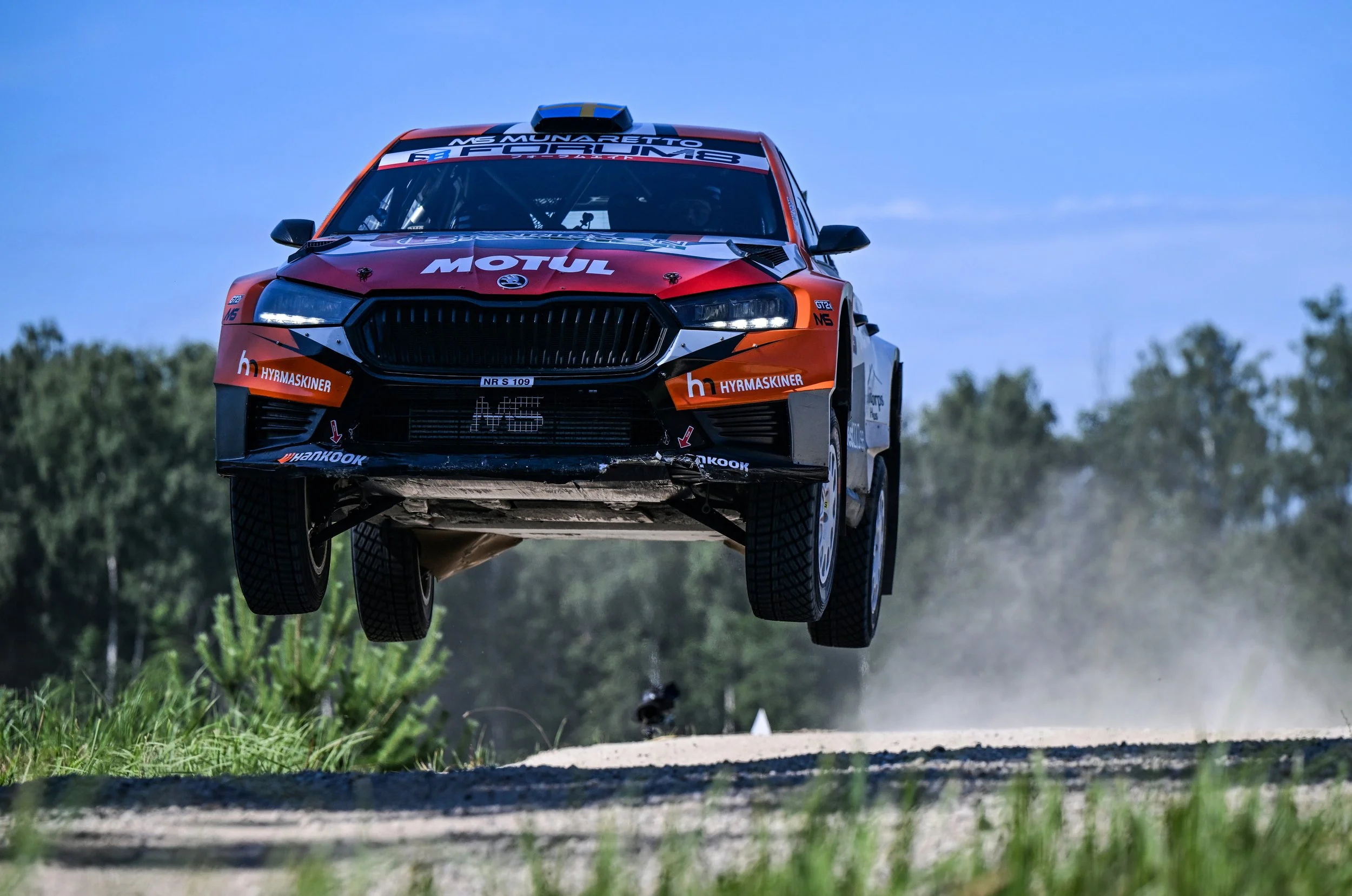 A rally car airborne above a gravel road with dust clouds, set against a green landscape and blue sky.