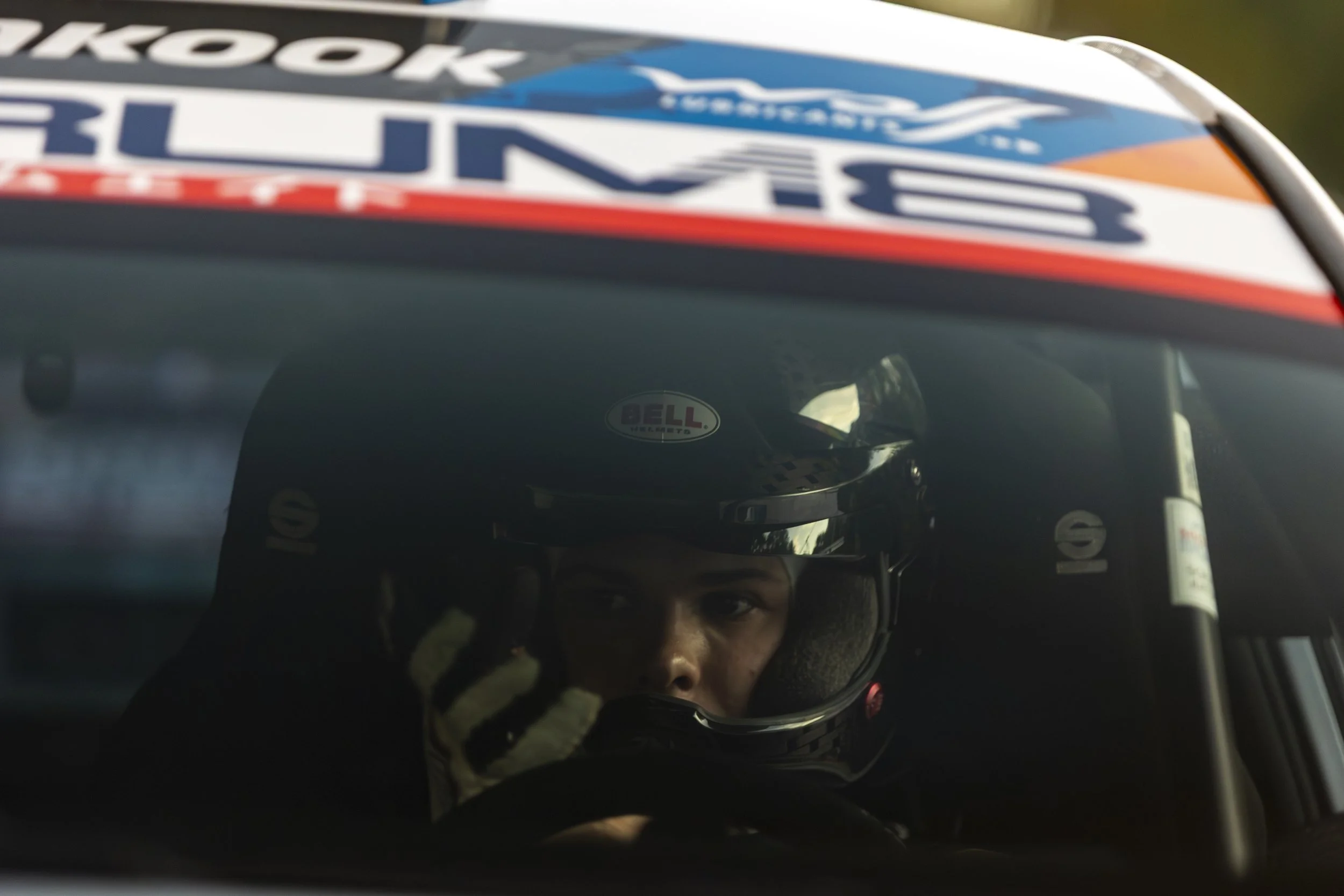 Close-up of a race car driver wearing a helmet, with logo and sponsor stickers, reflected in the helmet visor.