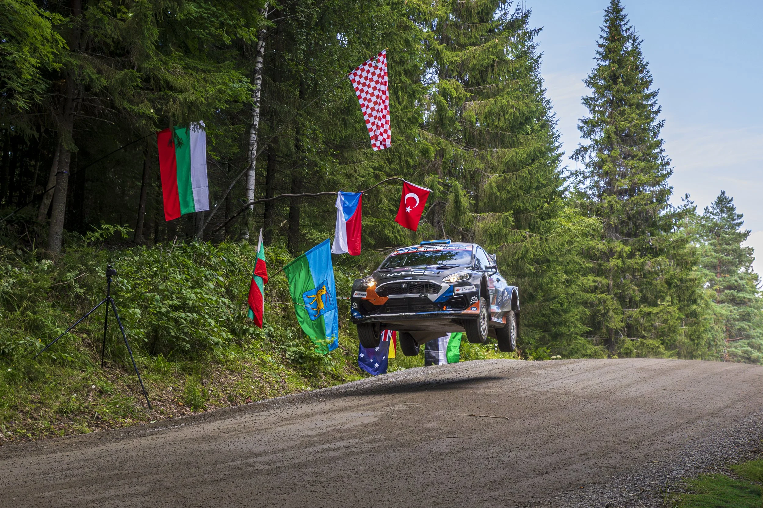 A rally car is airborne above a dirt road in a forested area, with flags from various countries hanging on a line above the road.