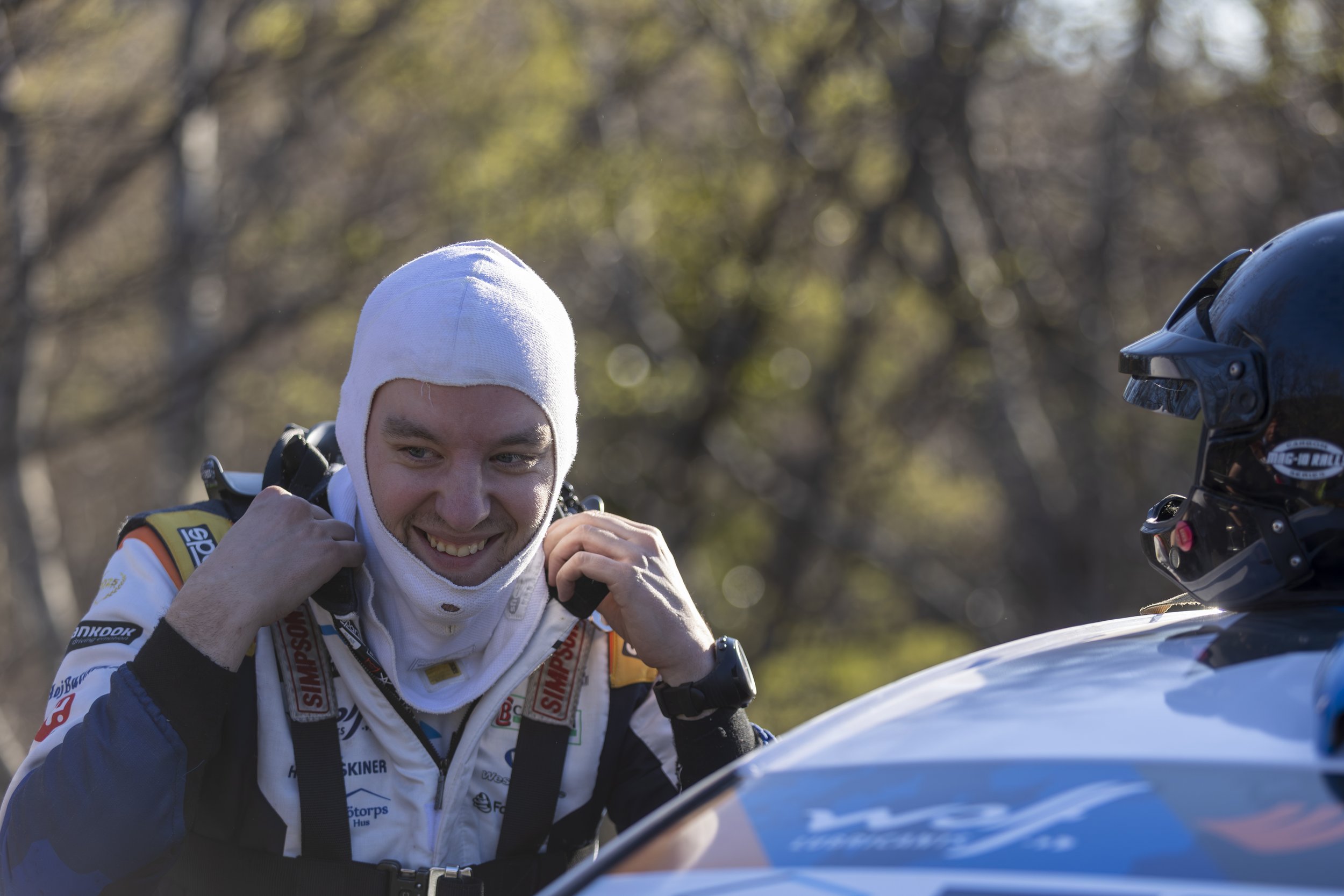 A race car driver wearing a white racing suit with patches and a white head sock, smiling and adjusting his helmet strap, standing next to a blue racing helmet, with a blurred outdoor background.