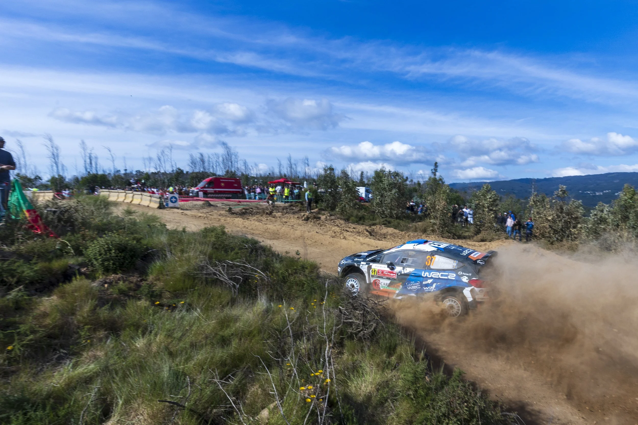 A rally car speeding on a dirt track, kicking up dust, with spectators and support vehicles in the background under a partly cloudy sky.