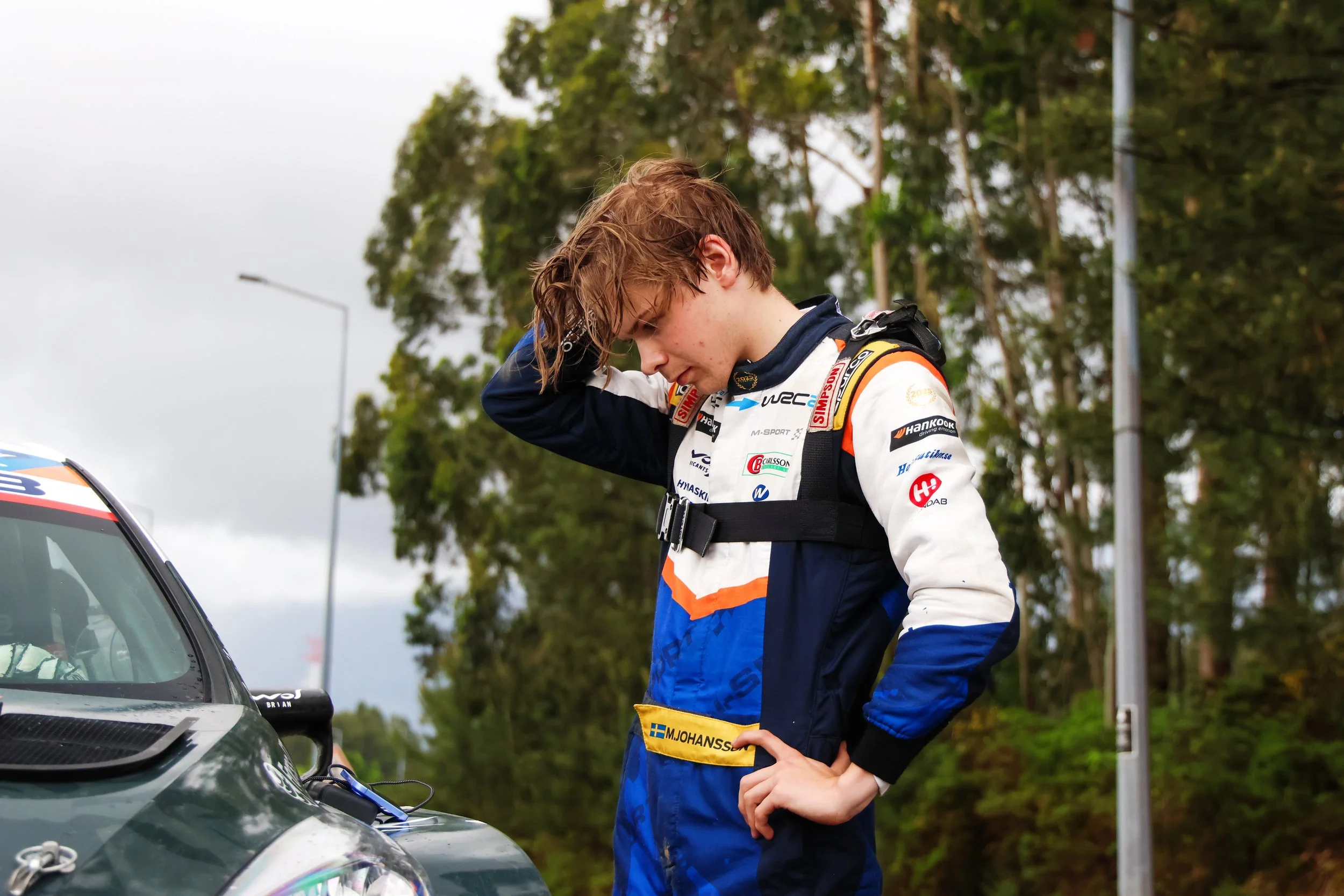 Young male race car driver in racing suit standing next to a race car, adjusting his helmet strap with trees and cloudy sky in the background.