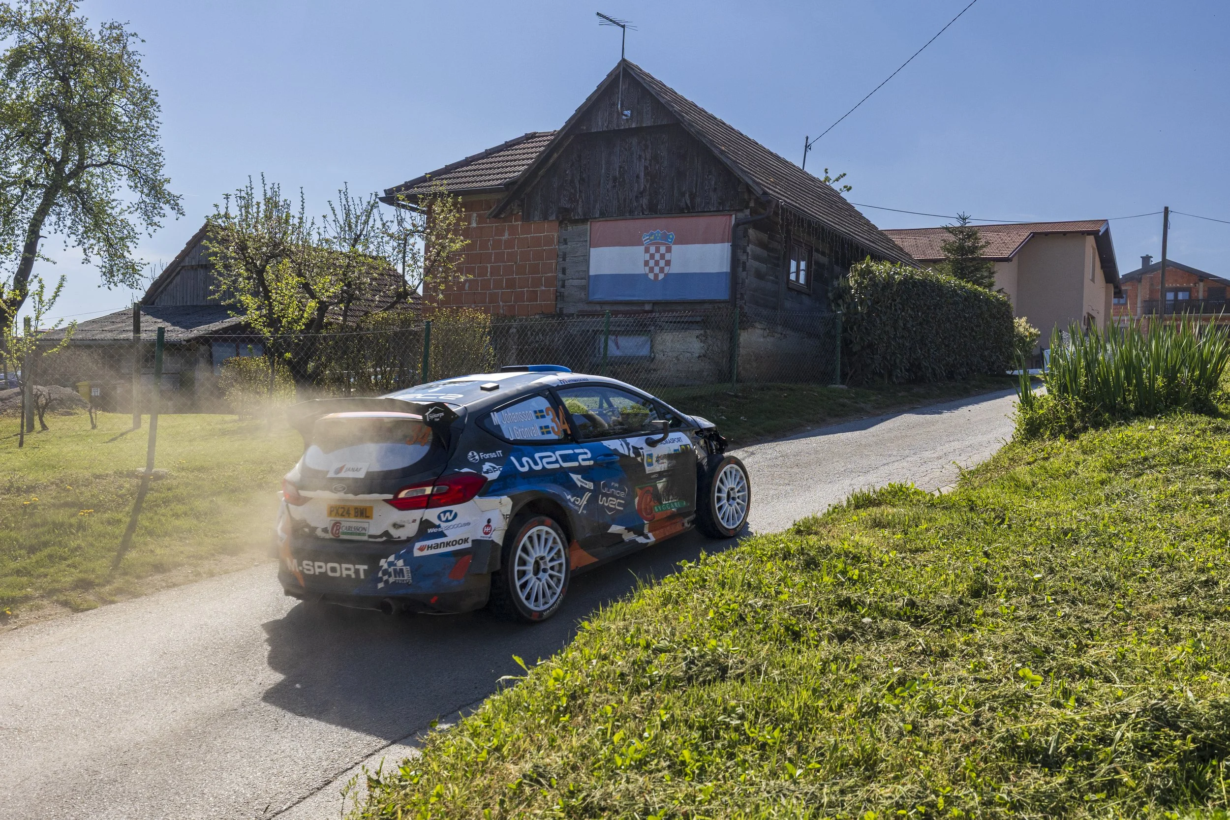 A racing rally car speeding along a rural road in front of houses, with a Croatian flag hanging on a house. The car has sponsorship decals and is kicking up dust as it drives.