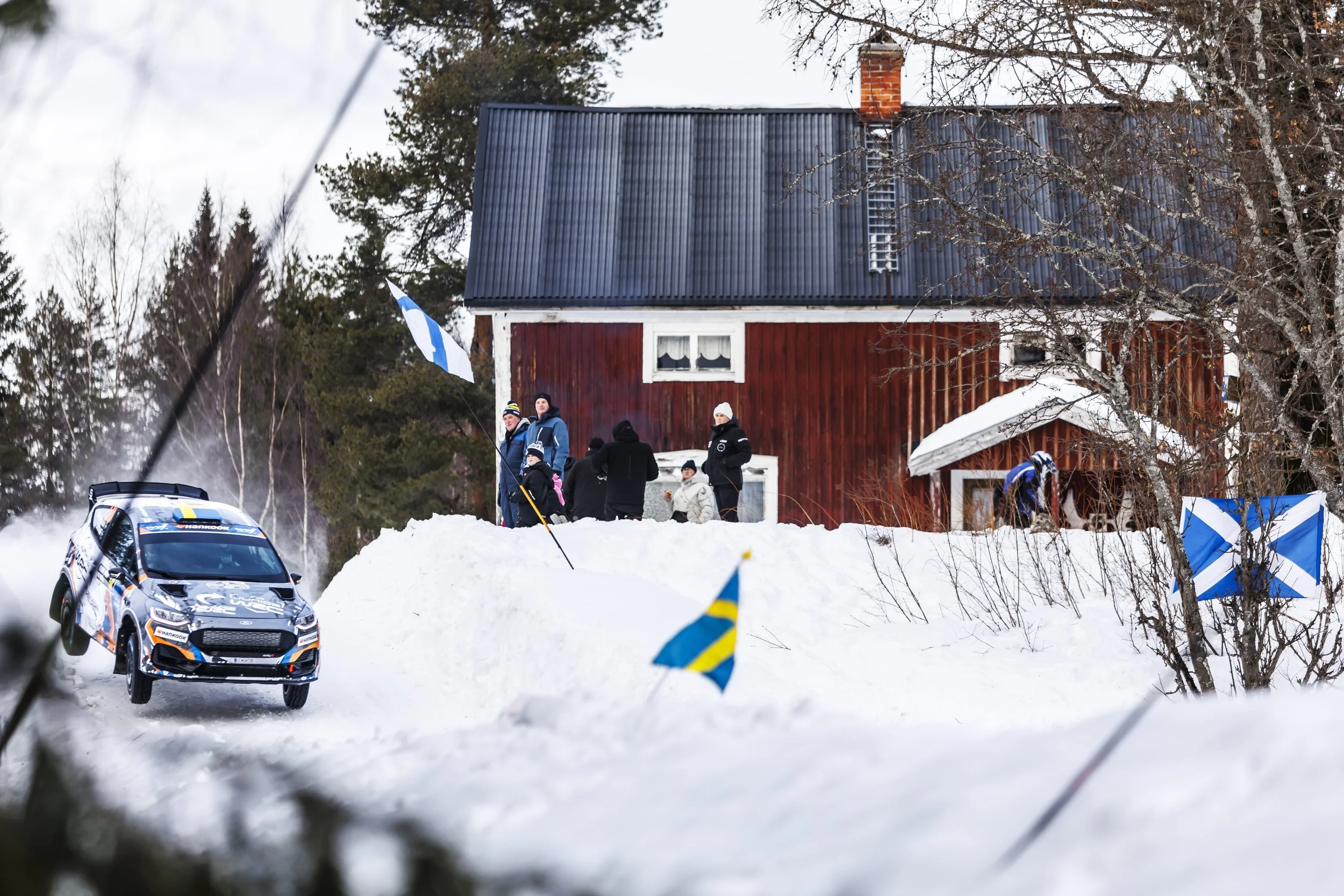 A rally race car on a snowy road with spectators watching beside a red house, and blue and white flags in the foreground.