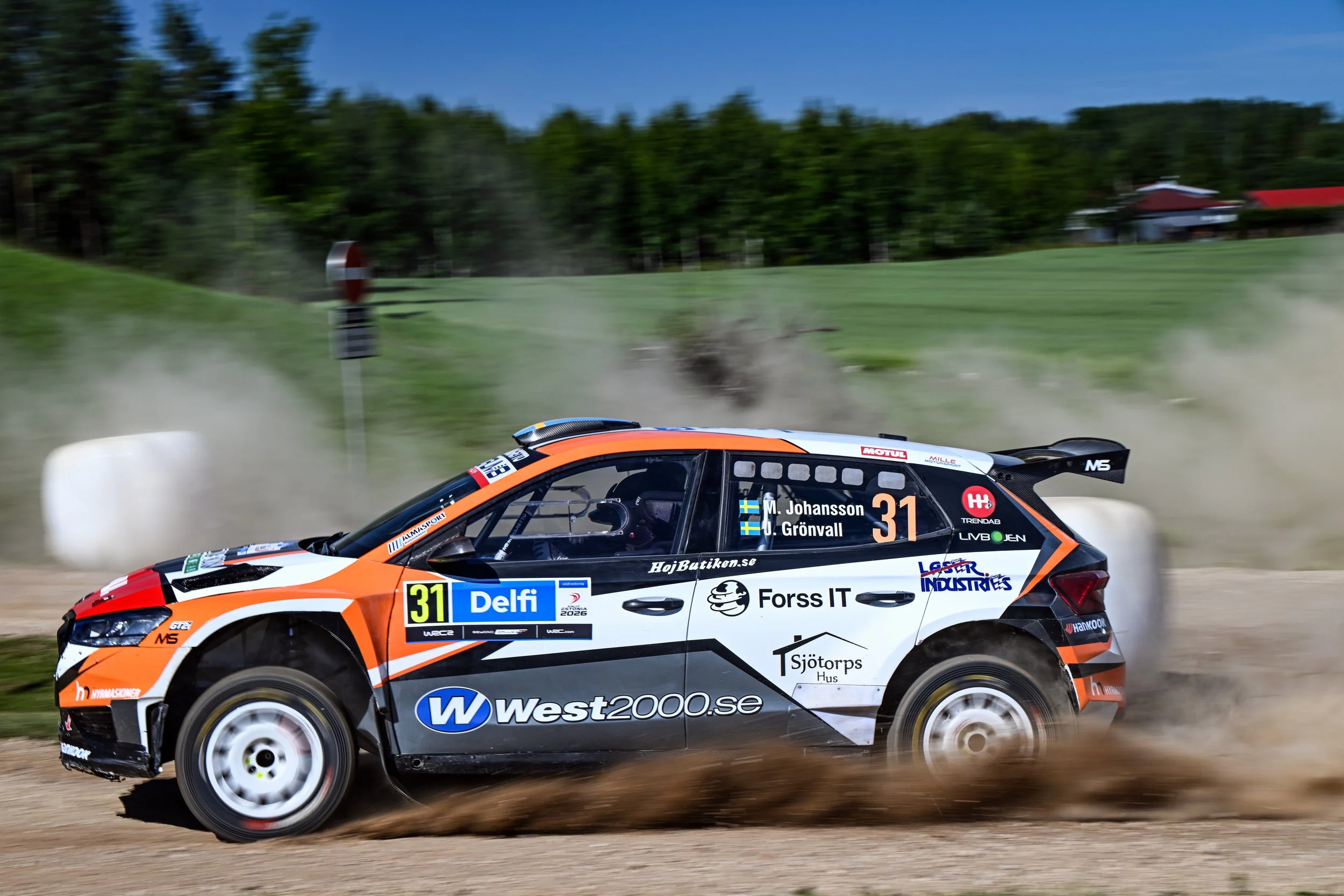 A rally car racing on a dirt road, kicking up dust, with a background of trees and a building with a red roof.