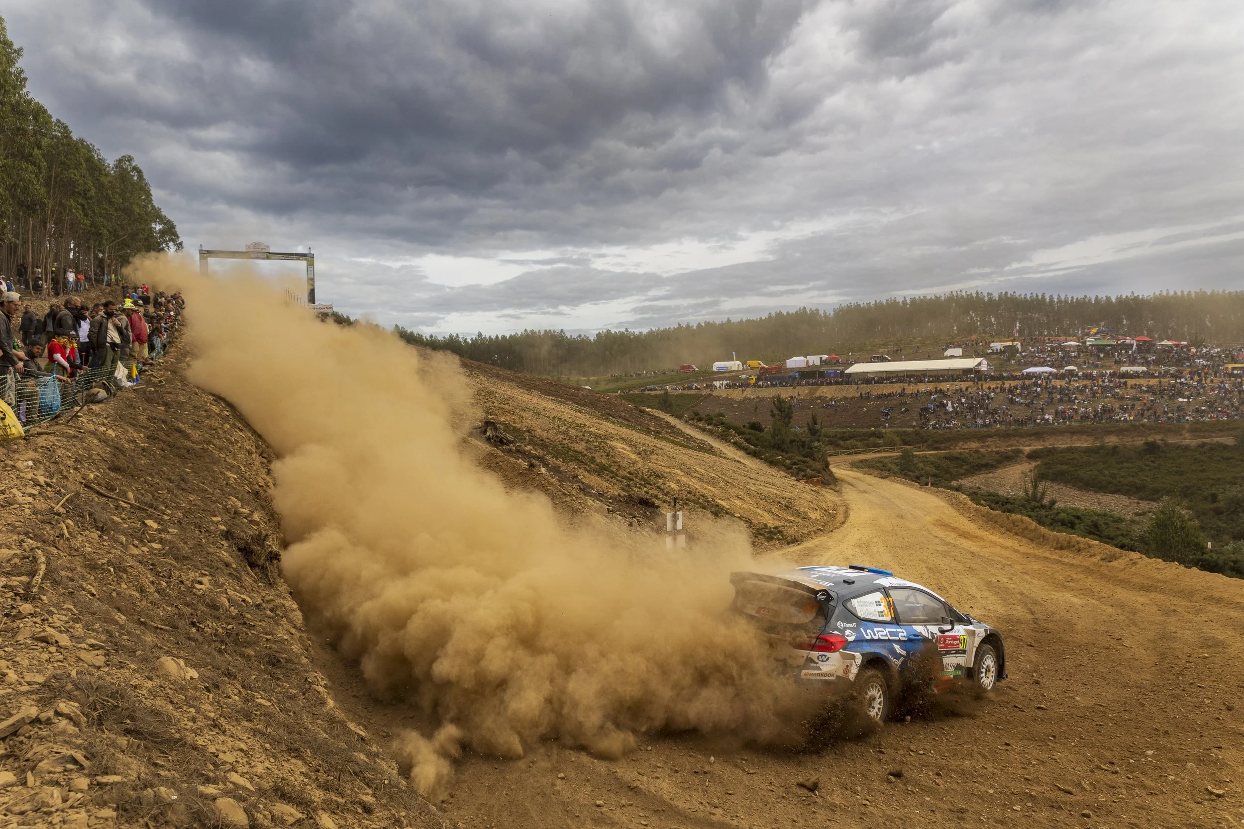 A rally car race on a dirt track, with a car kicking up dust, spectators watching from the hillside, and tents and vehicles in the background under cloudy skies.