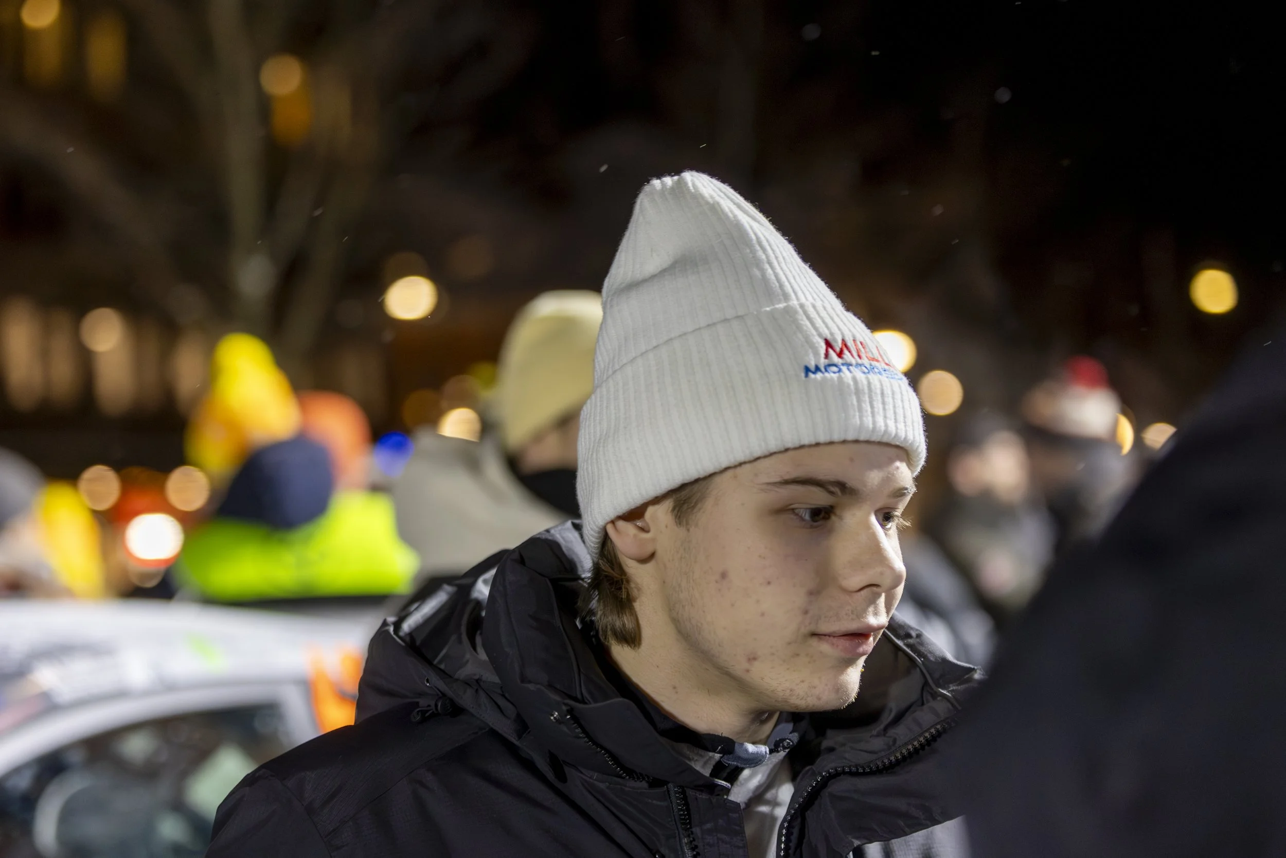 A young person wearing a white knit hat with the logo 'Milwaukee Motors' and a black jacket, outdoors at night with blurred lights and people in the background.