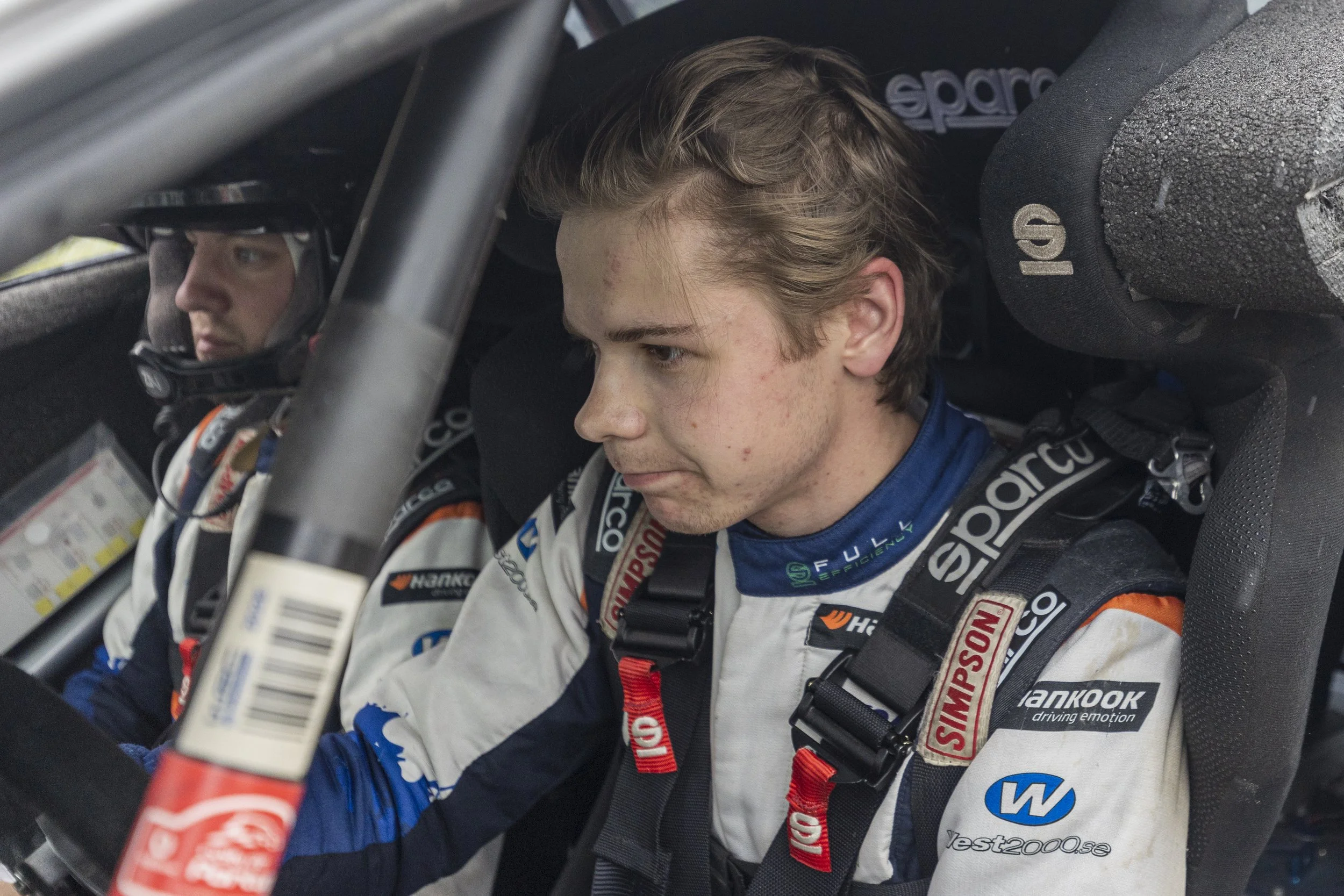 Two race car drivers sitting inside a race car, wearing racing suits and helmets, preparing for a race.