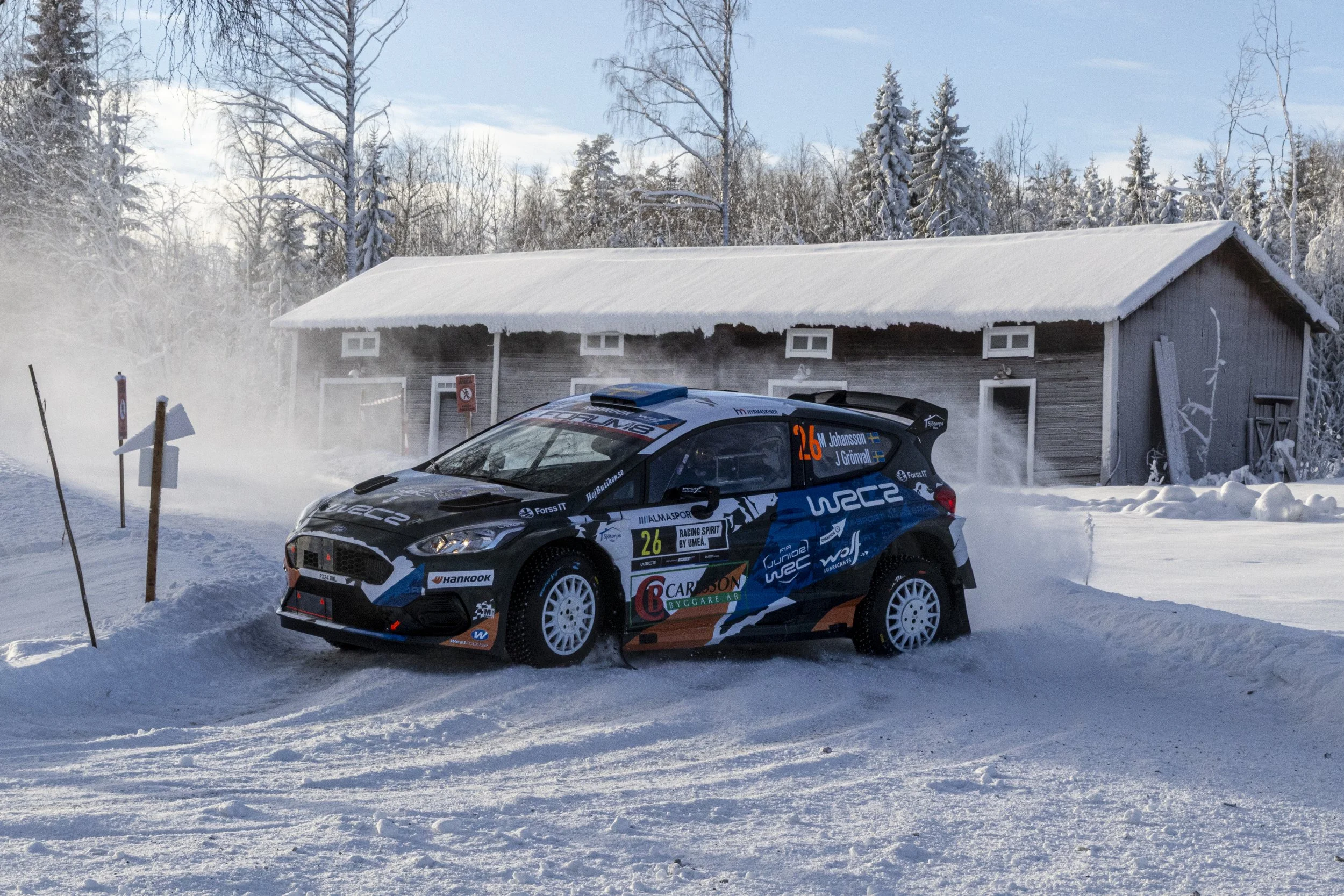 A rally car on a snow-covered ground near a wooden building and snow-covered trees, with a blue sky overhead.