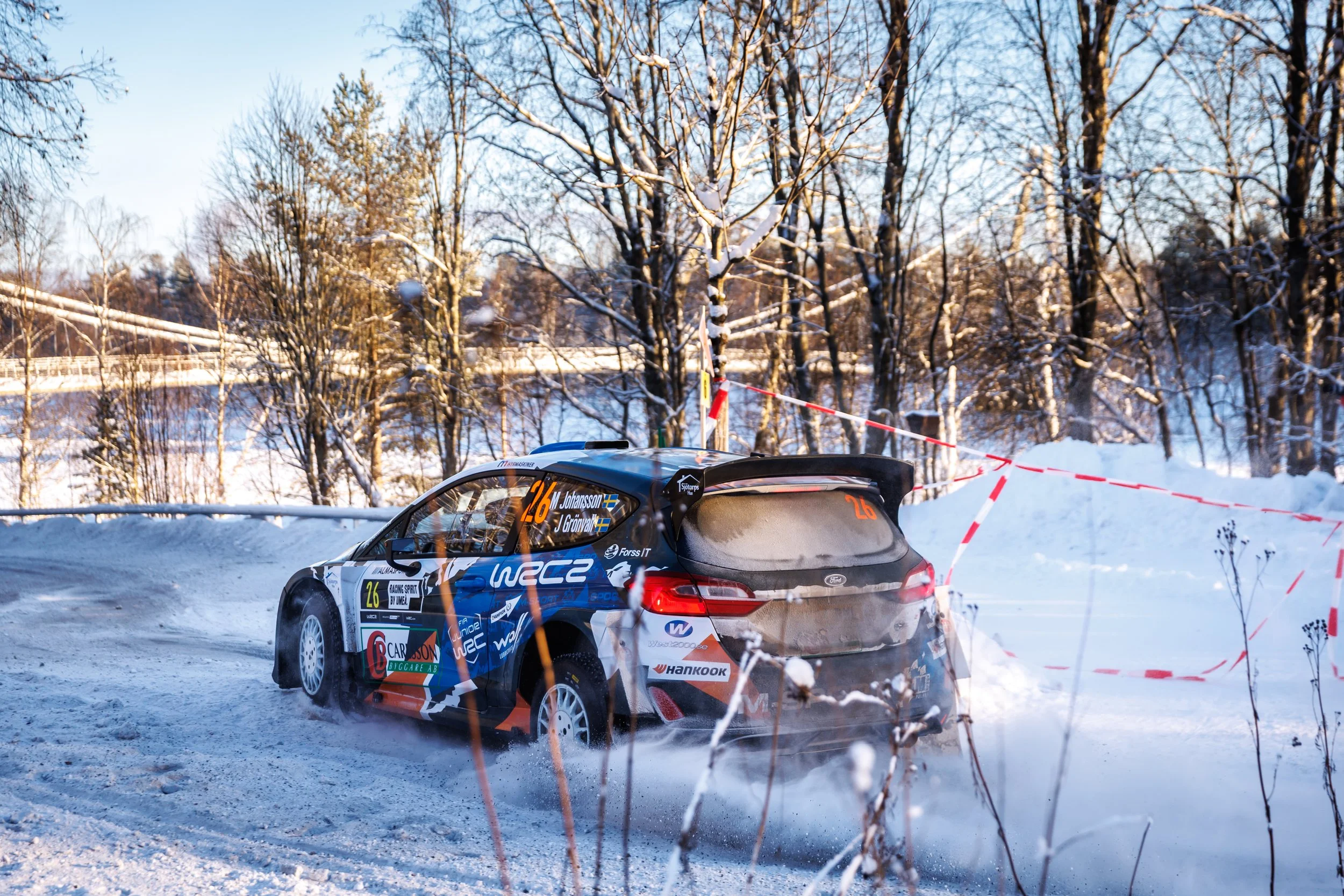 A rally car driving on a snowy road in a winter landscape with snow-covered trees and a clear blue sky.