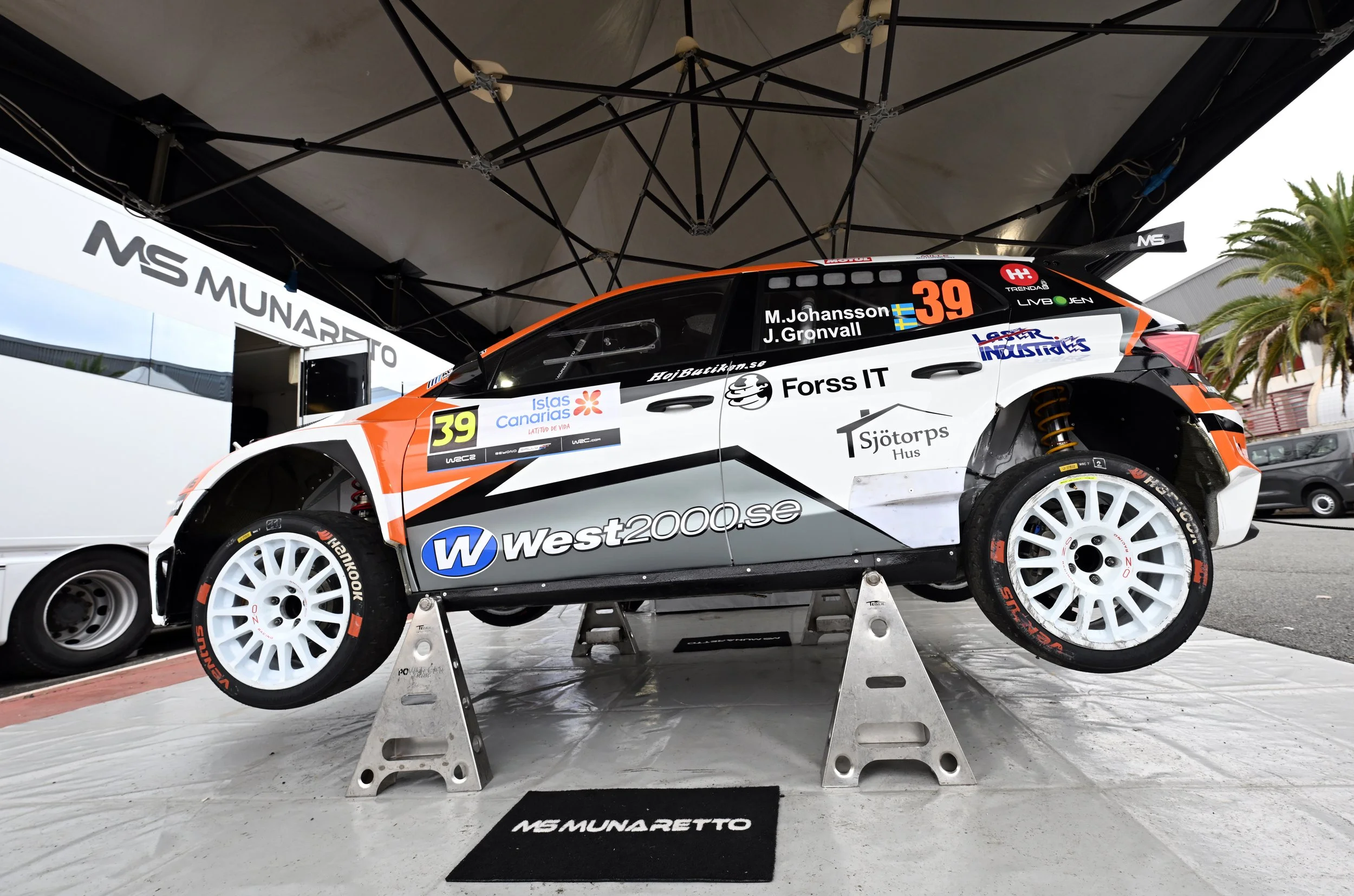 A rally car with the number 39 and the names M. Johansson and J. Gronvall on the side, parked on a stand under a canopy with various sponsor logos. The car is black, white, orange, and red, and is elevated on two jack stands.