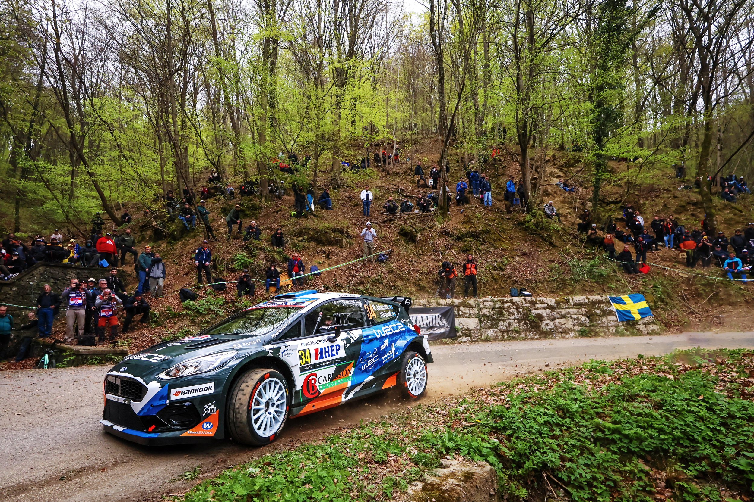 A black and blue rally car racing on a dirt road with a crowd of spectators standing on a hillside in the background. The spectators are sitting and standing among trees and some hold flags, one of which is Swedish. The scene is in a wooded area with