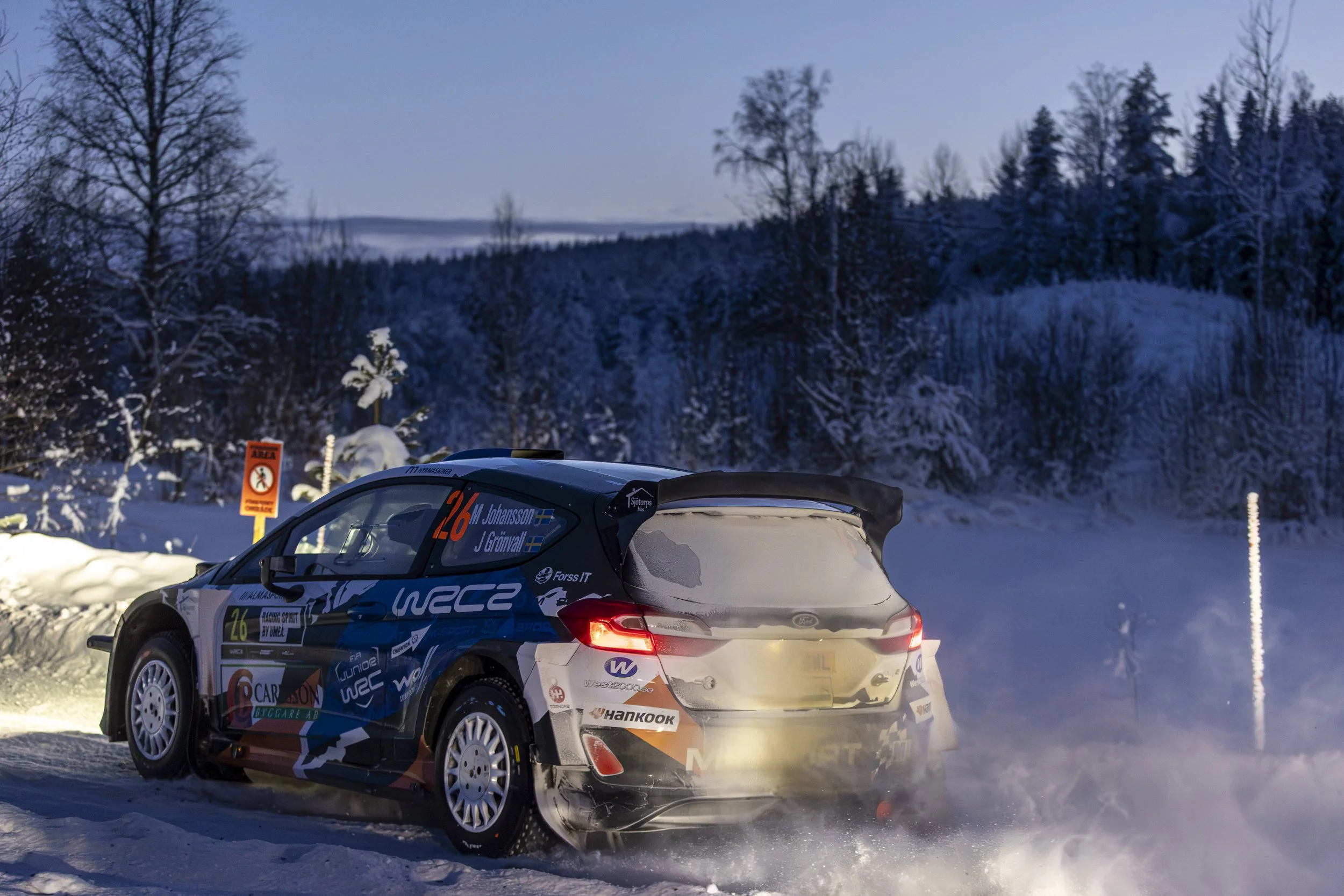 A rally car speeding on a snowy mountain road, with snow-covered trees and a blue sky in the background.