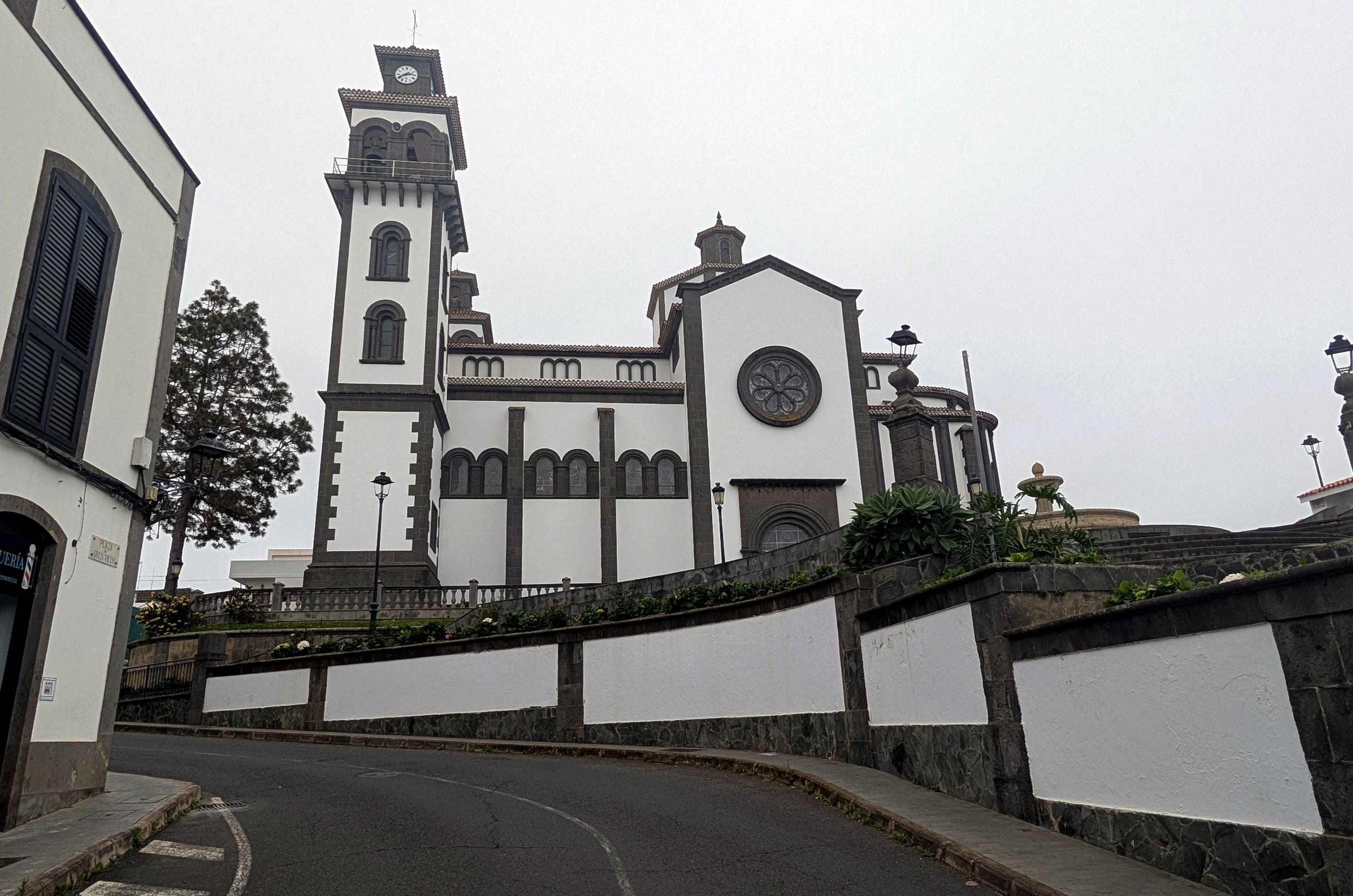 A large white church with dark trim, multiple towers, arched windows, a circular stained glass window, set on a hillside with stone railings, lamp posts, and trees under an overcast sky.