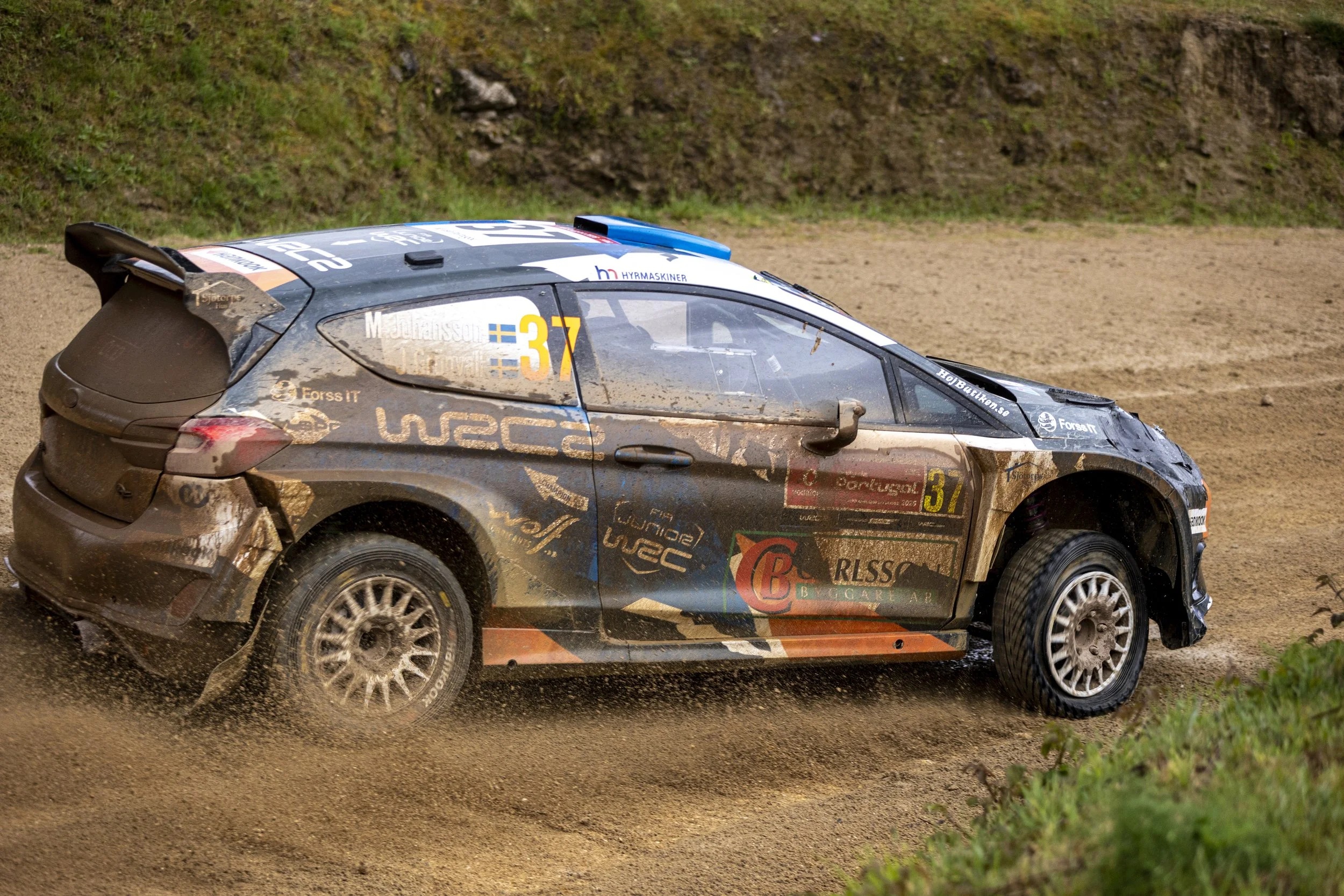 A rally car speeding on a dirt track kicking up mud, with a background of grass and a hillside.