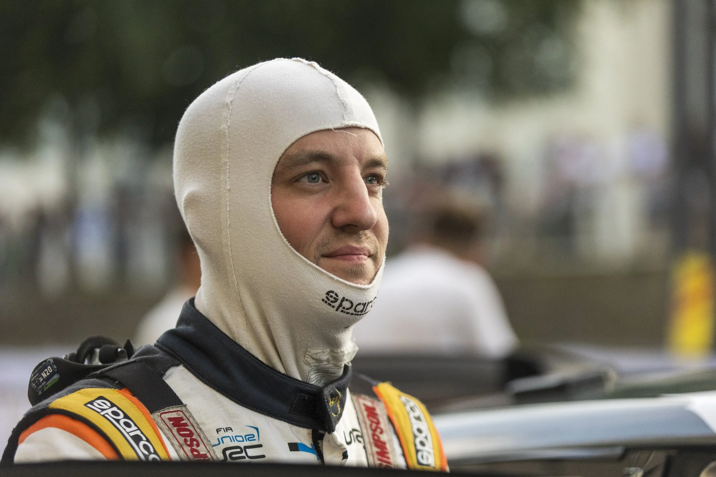 A race car driver wearing a white racing helmet and a racing suit with sponsor patches, standing outdoors with a blurred background of trees and people.