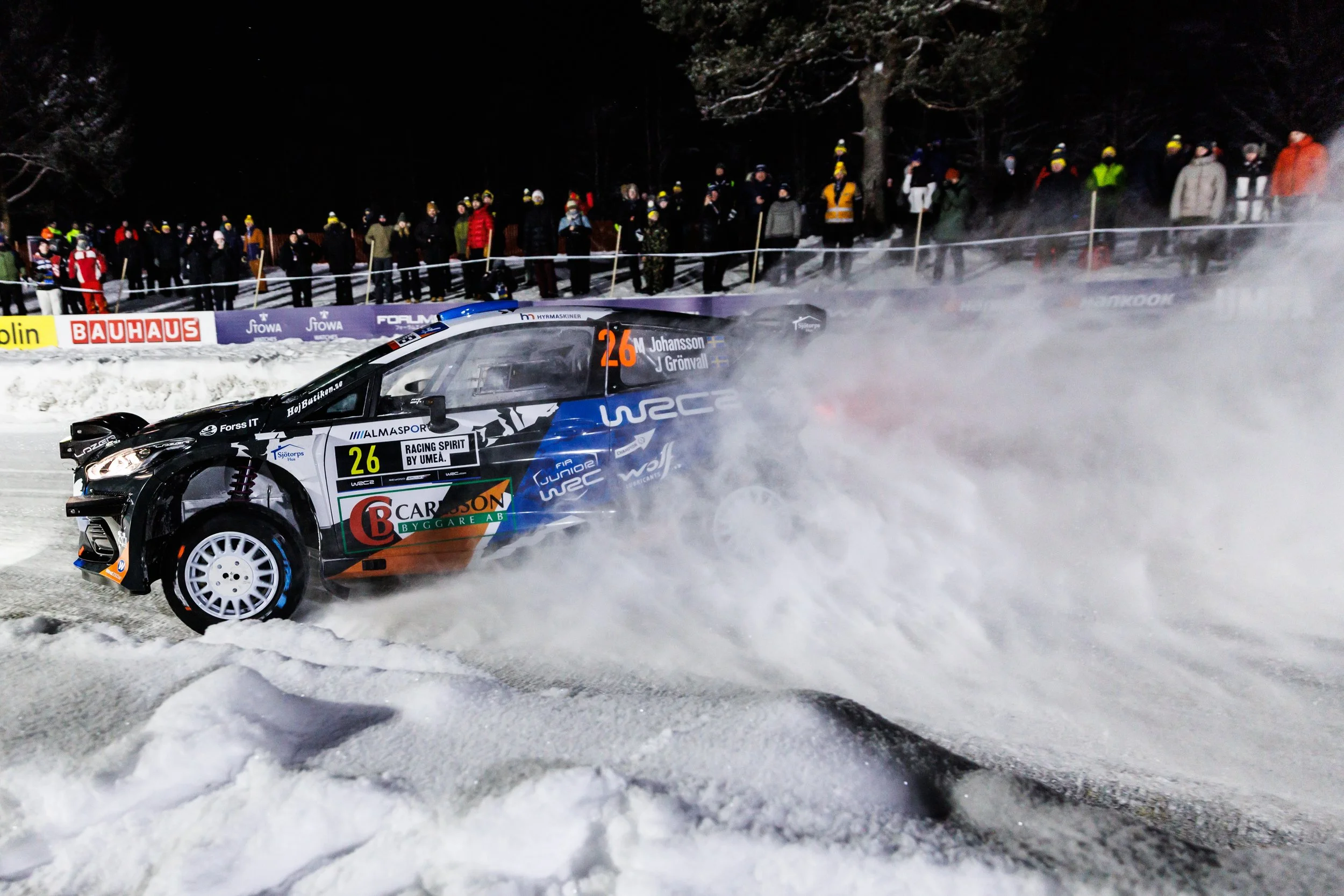 A rally car racing on snow at night, kicking up snow and smoke, with spectators watching from beyond a barrier.