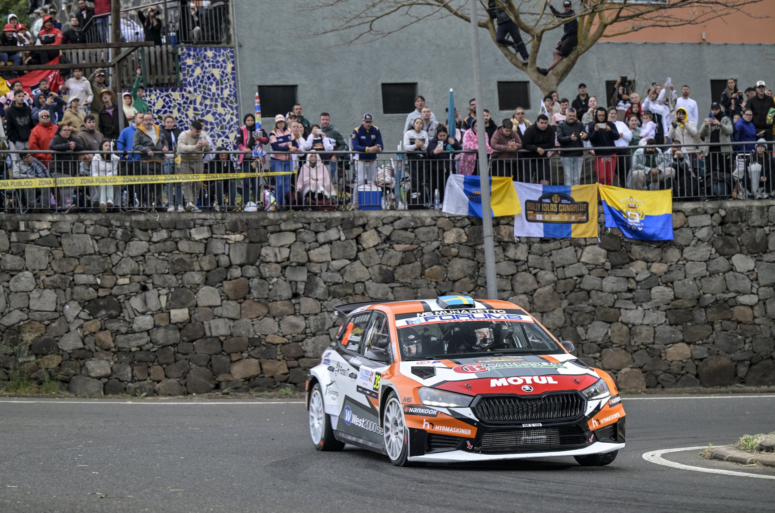 A rally car racing on a road with spectators watching from behind a fence and a stone wall, some taking photos. The car has a black, white, orange, and green livery, with sponsor logos.