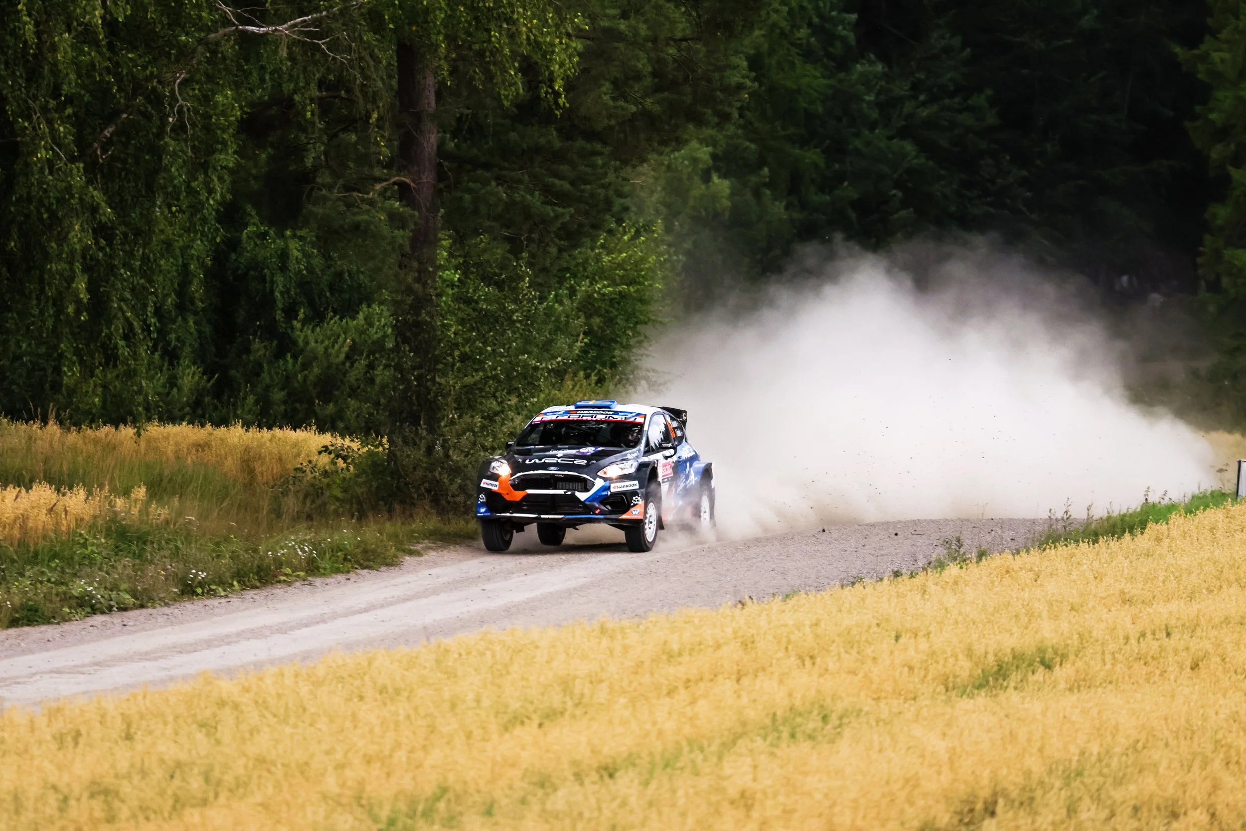 A rally car driving on a dirt road through a forested area with tall trees and green foliage, kicking up dust behind it.