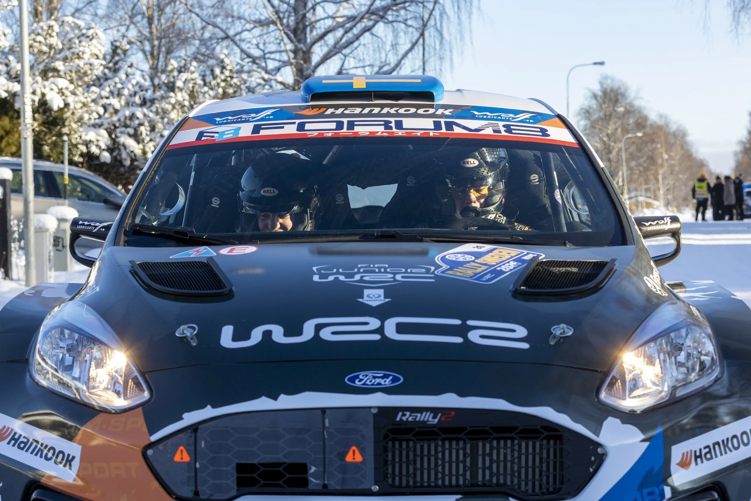 A black rally car with various sponsor logos and decals, including Ford, Hankook, and WRC, is on a snowy road with two drivers wearing helmets visible through the windshield. Snow-covered trees and a group of people are in the background.