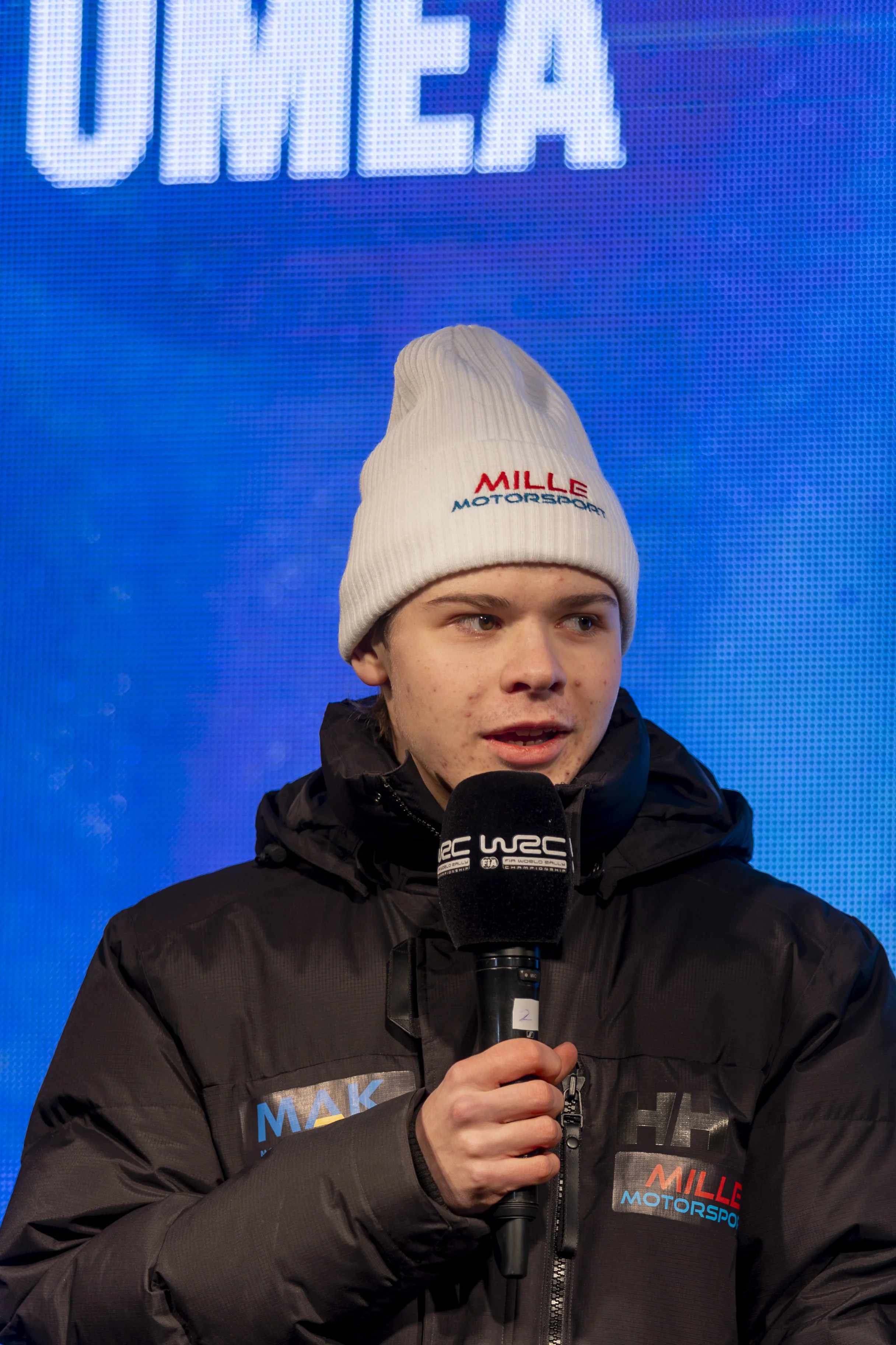 Young male journalist with brown hair and fair skin wearing a white beanie hat with red and blue logo, black jacket with logos, holding a microphone with event branding, standing in front of a blue LED screen showing the word 'UEA' in large illuminat