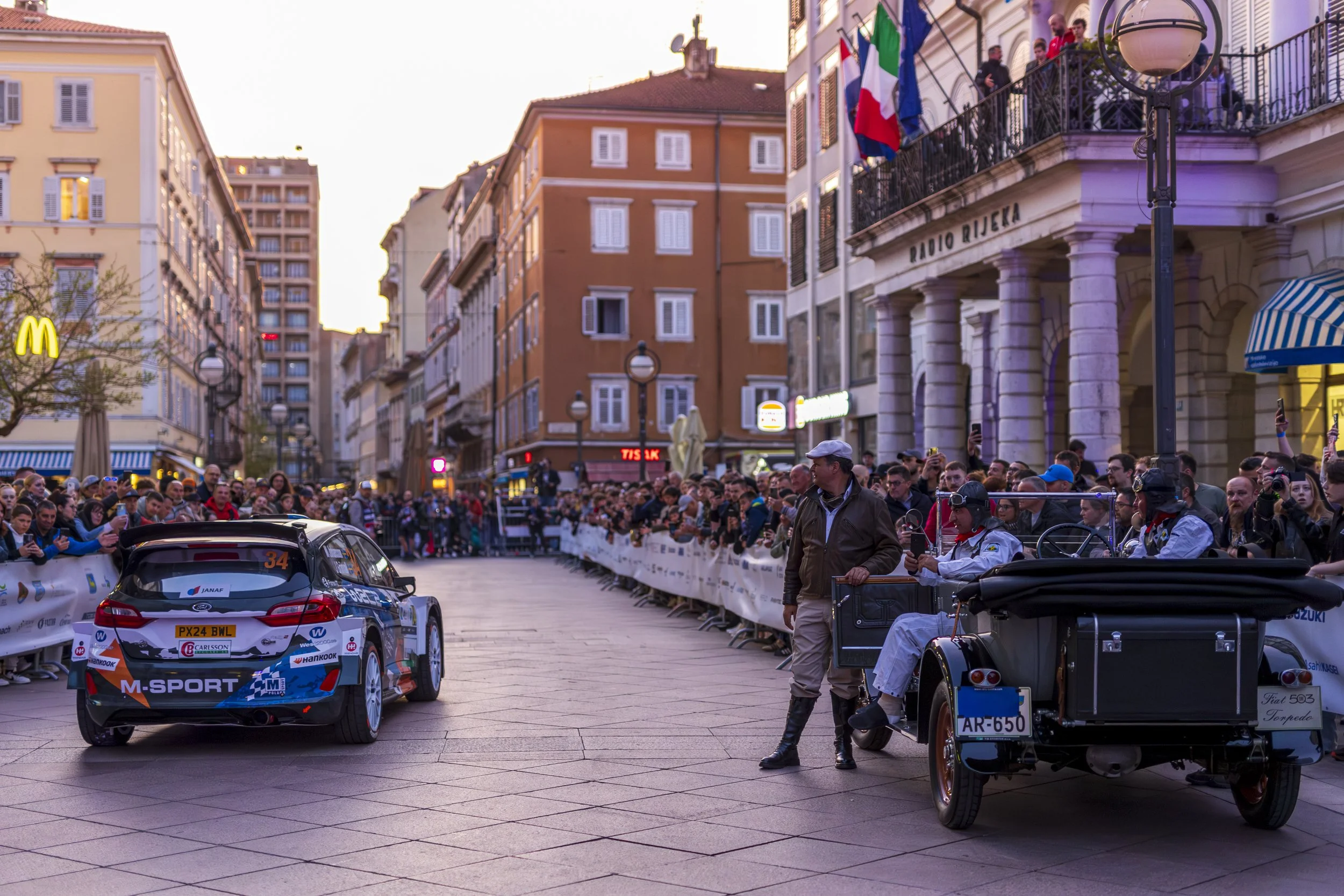 A race car with sponsor logos on the rear is driving through a street lined with spectators on both sides, behind barriers. People are taking photos and watching the event. Buildings with flags and signs are visible in the background, and the sky is 