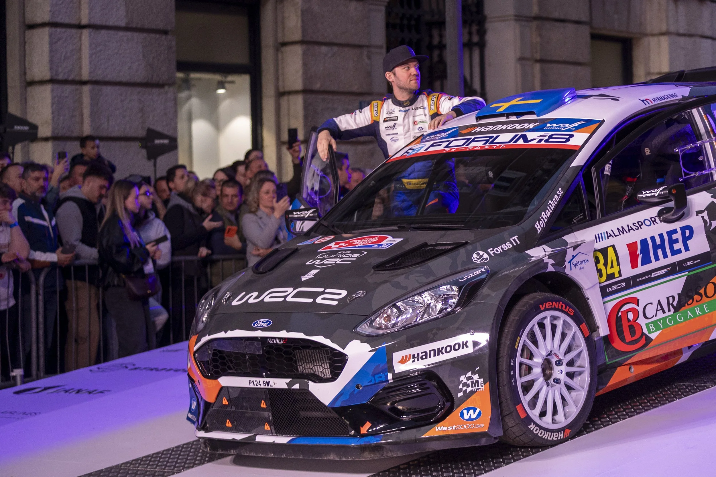 A rally car on display with a man leaning on it, surrounded by a crowd of spectators, in an urban setting at night.