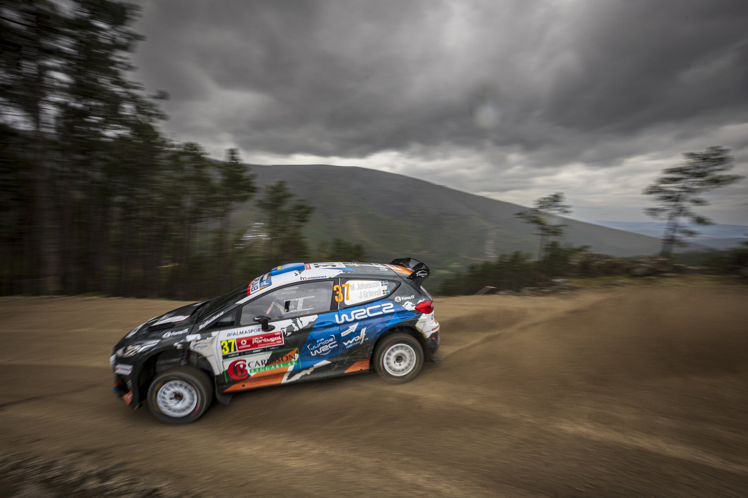 A rally car racing on a dirt road with a mountainous landscape and dark, cloudy sky in the background.