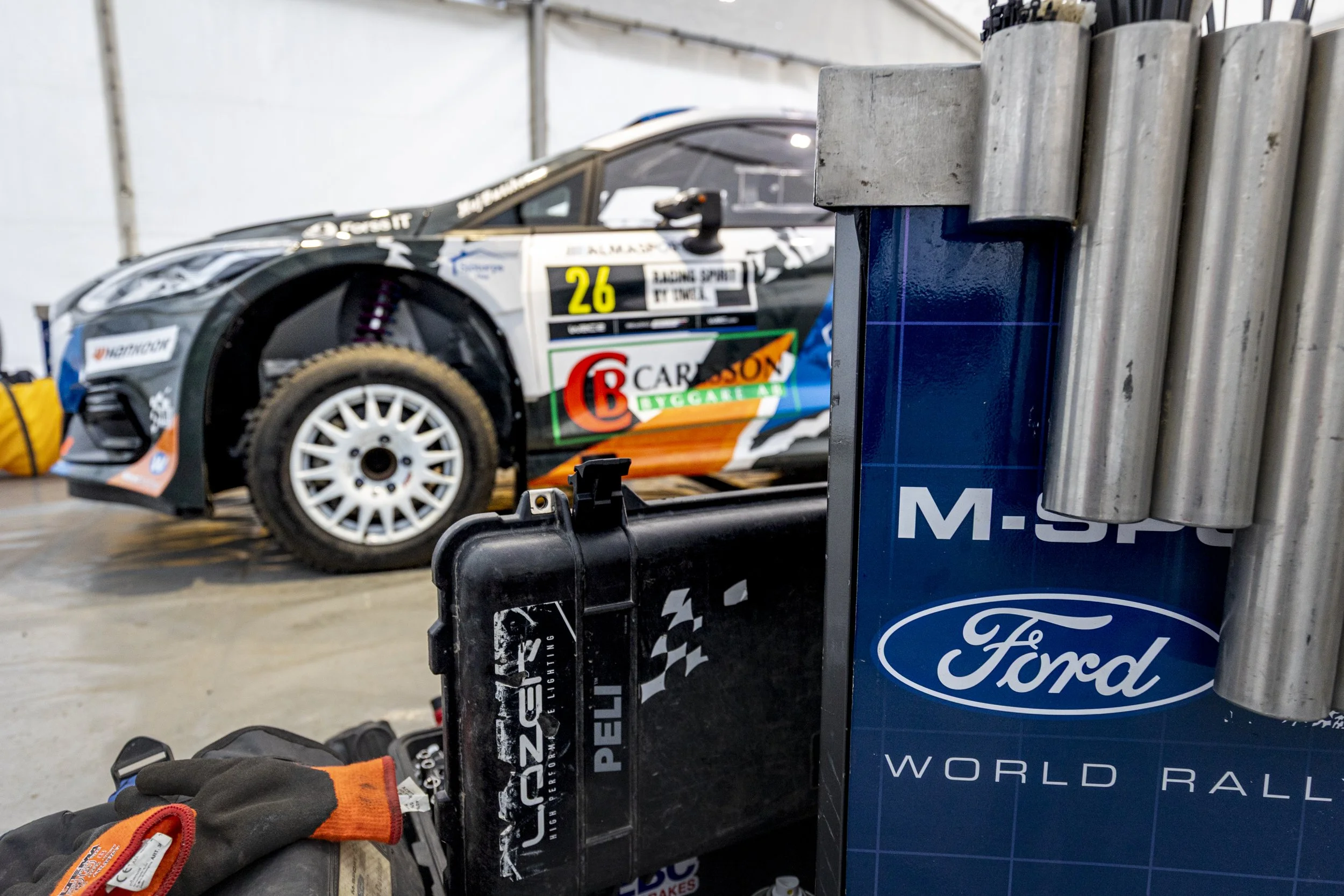 A race car with sponsorship decals and the number 26 is parked in a garage. There are racing gloves and equipment in the foreground.