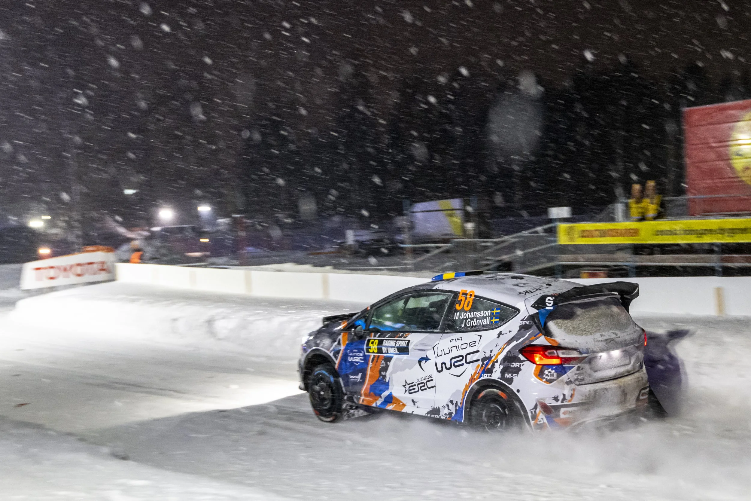 A rally car racing on snow-covered ground in snowy weather at night, with snow falling and stadium lights illuminating the scene.