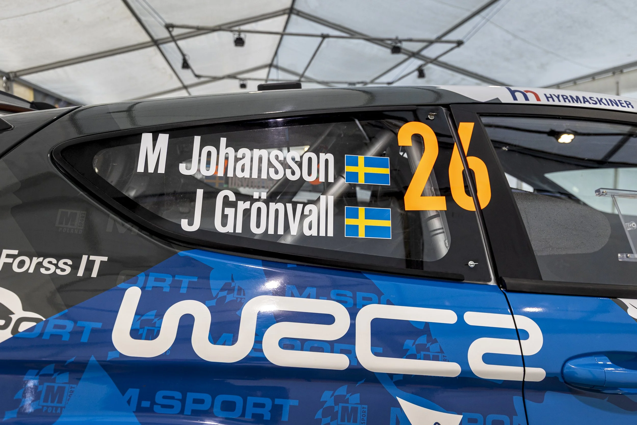 Close-up of a rally car with driver and co-driver names 'M. Johansson' and 'J. Grönyall' on the side window, along with Swedish flags, and the number 26, under a large white tent.