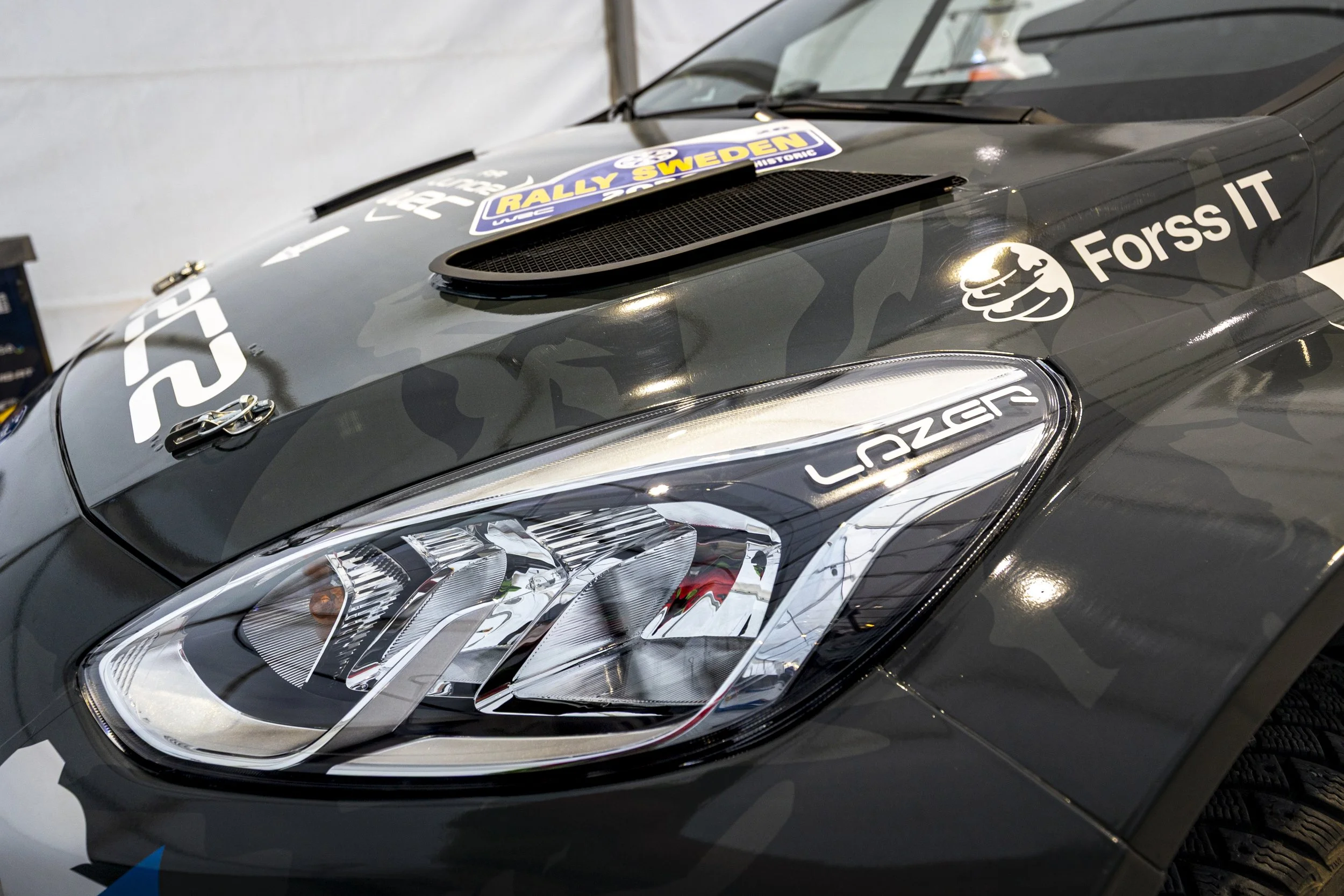 Close-up of a black race car with racing decals and a headlight, featuring a sticker that reads 'RALLY SWEDEN' on the hood.
