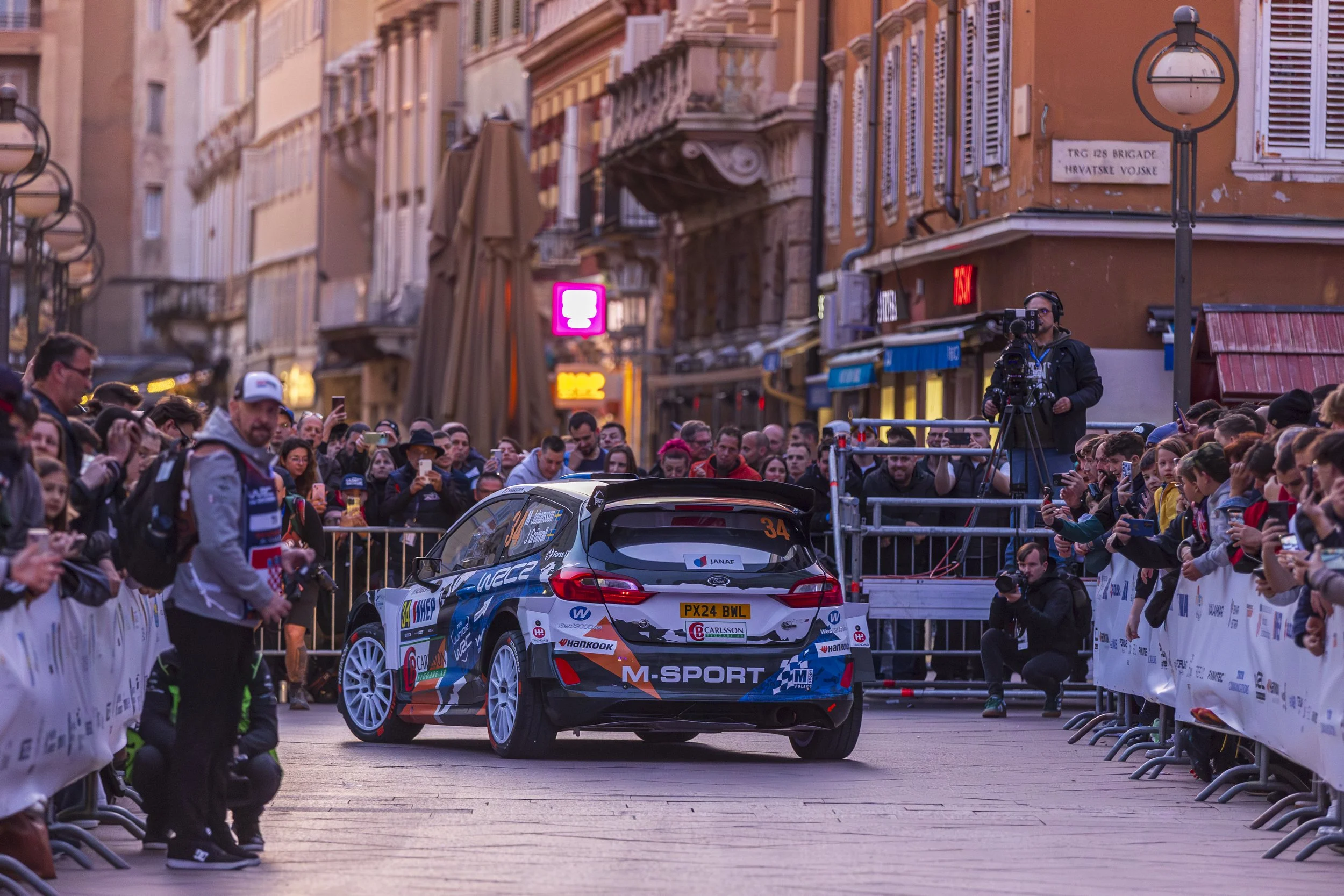 A rally car racing through a street with spectators on both sides, photographers, and a cameraman recording. The buildings are European in style, with balconies and shuttered windows. The car has sponsorship decals and the number 34, and the crowd ap
