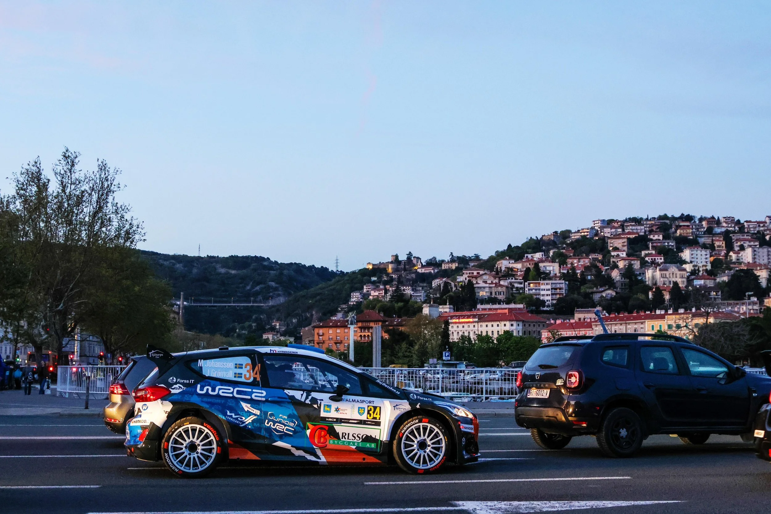 A rally car with number 34 and sponsor logos parked on a city street, with a hillside residential area and blue sky in the background.