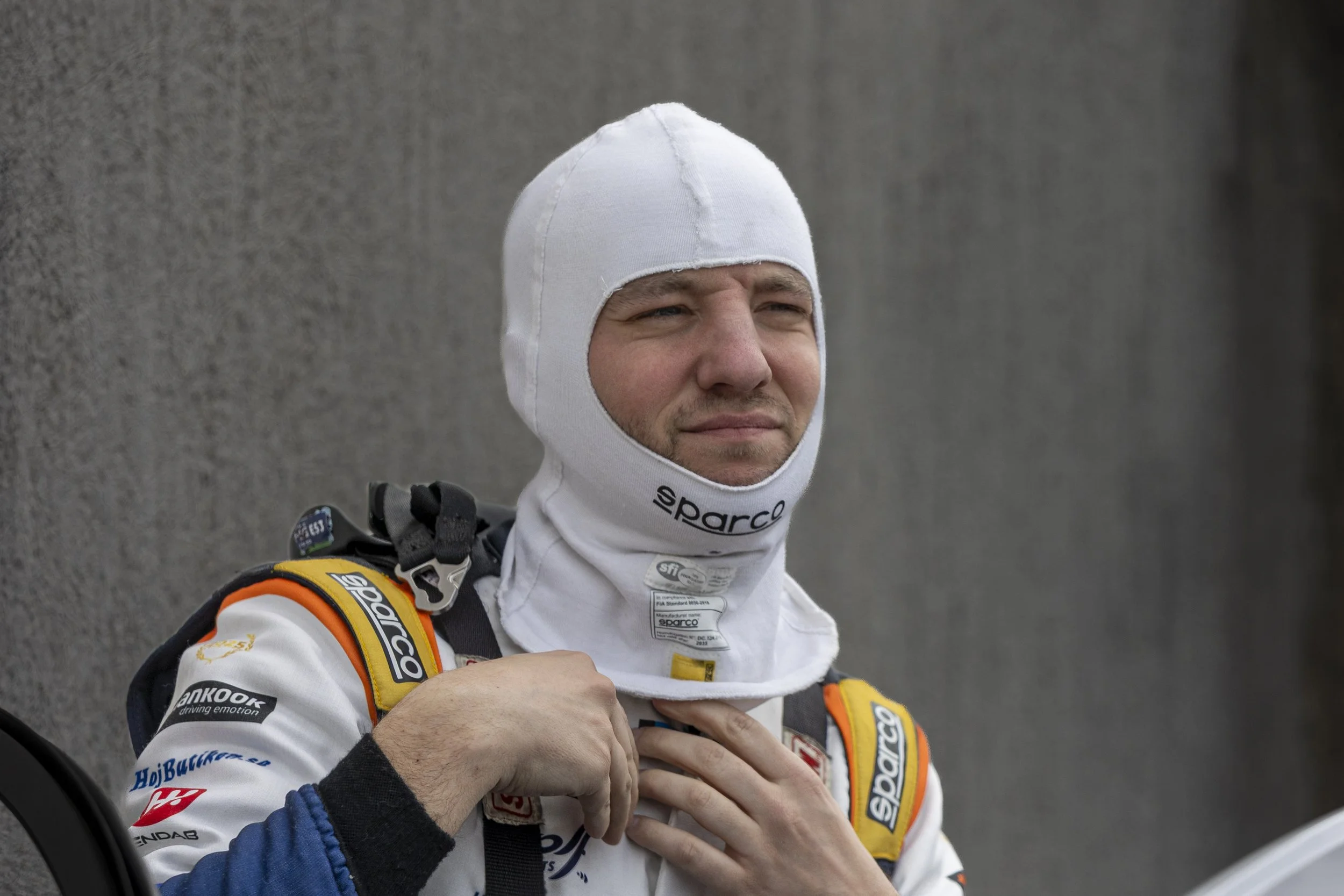 A race car driver in a white racing helmet and suit with sponsor logos, adjusting his collar while standing against a gray background.
