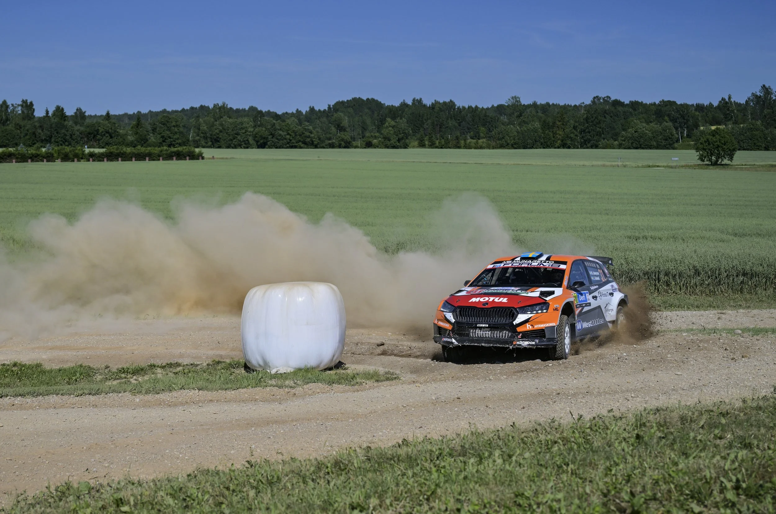 Rally car kicking up dust on a dirt track, with a large hay bale in the foreground and green fields, trees, and a blue sky with few clouds in the background.