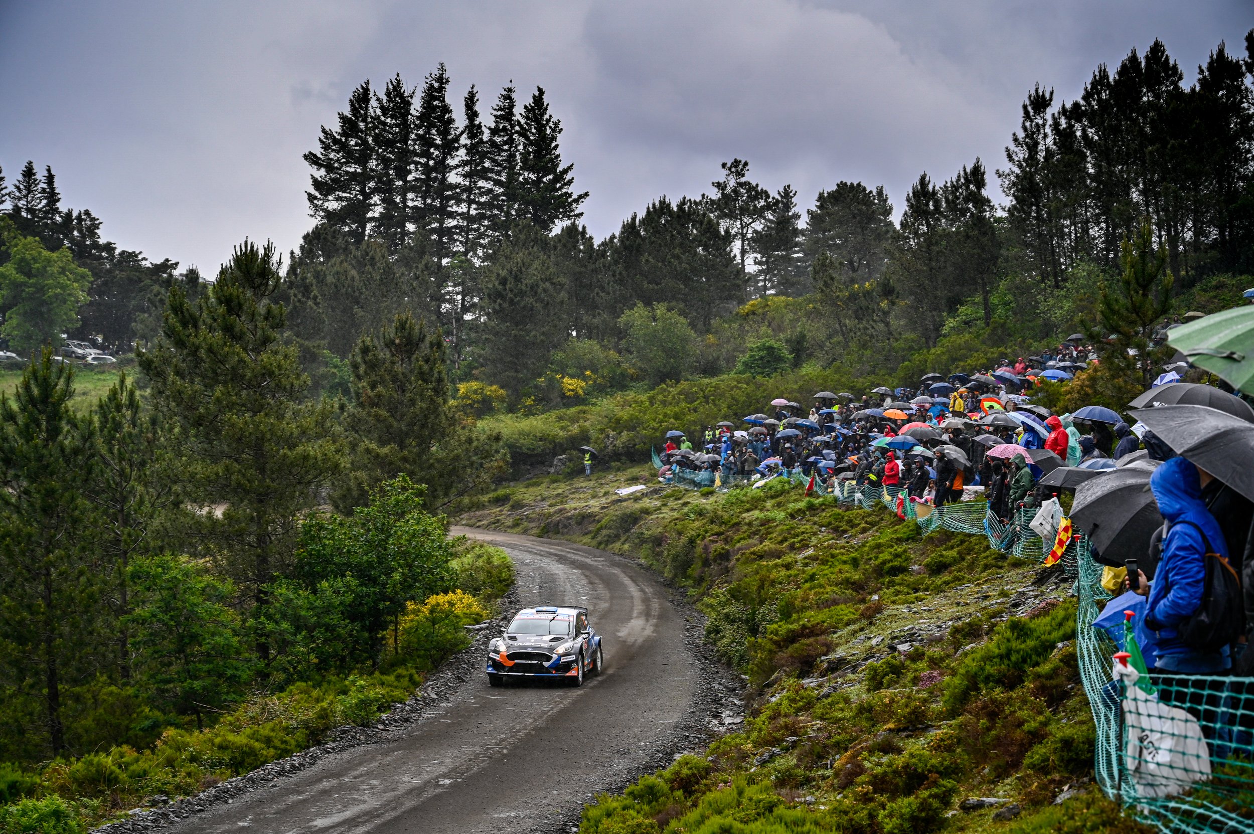 A racing car on a dirt rally stage with spectators watching and holding umbrellas on a rainy day.