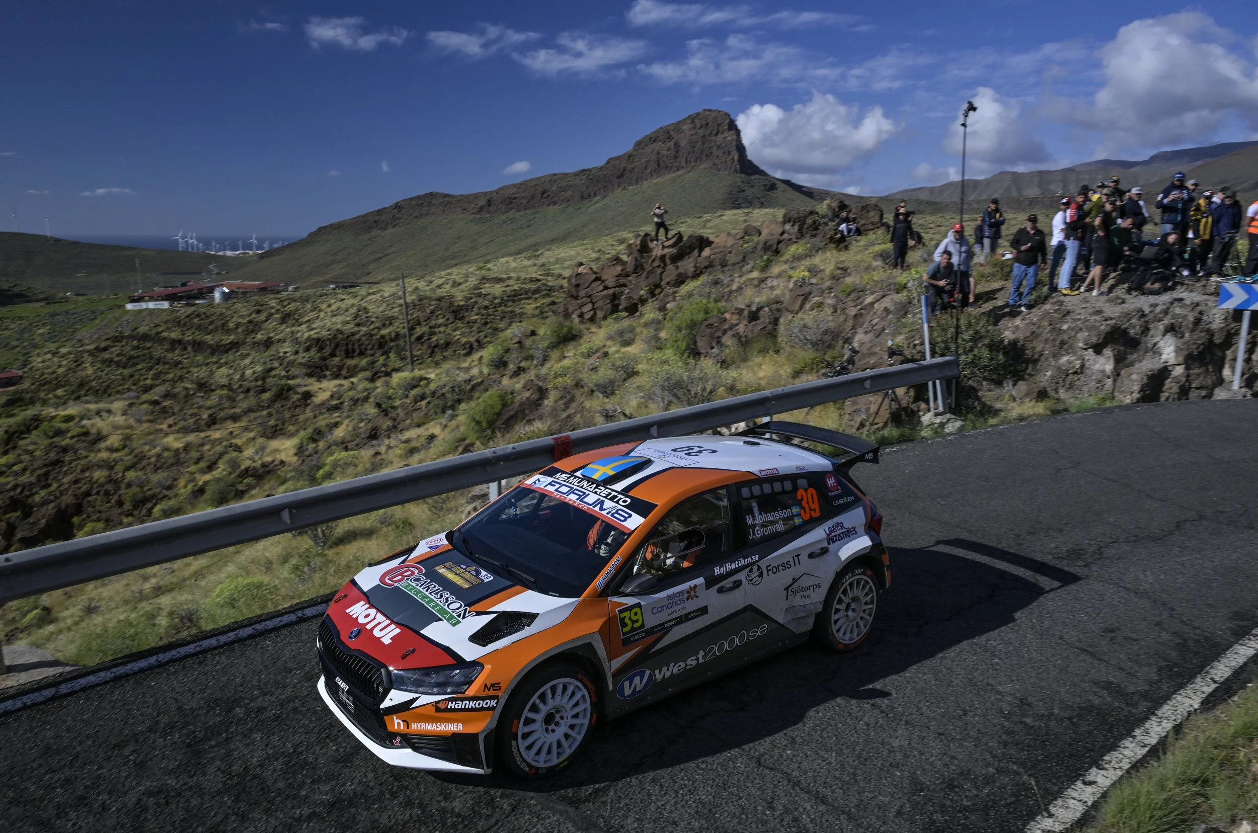 A rally car racing on a mountain road with spectators watching from the side, and wind turbines visible in the distant background, under a partly cloudy sky.