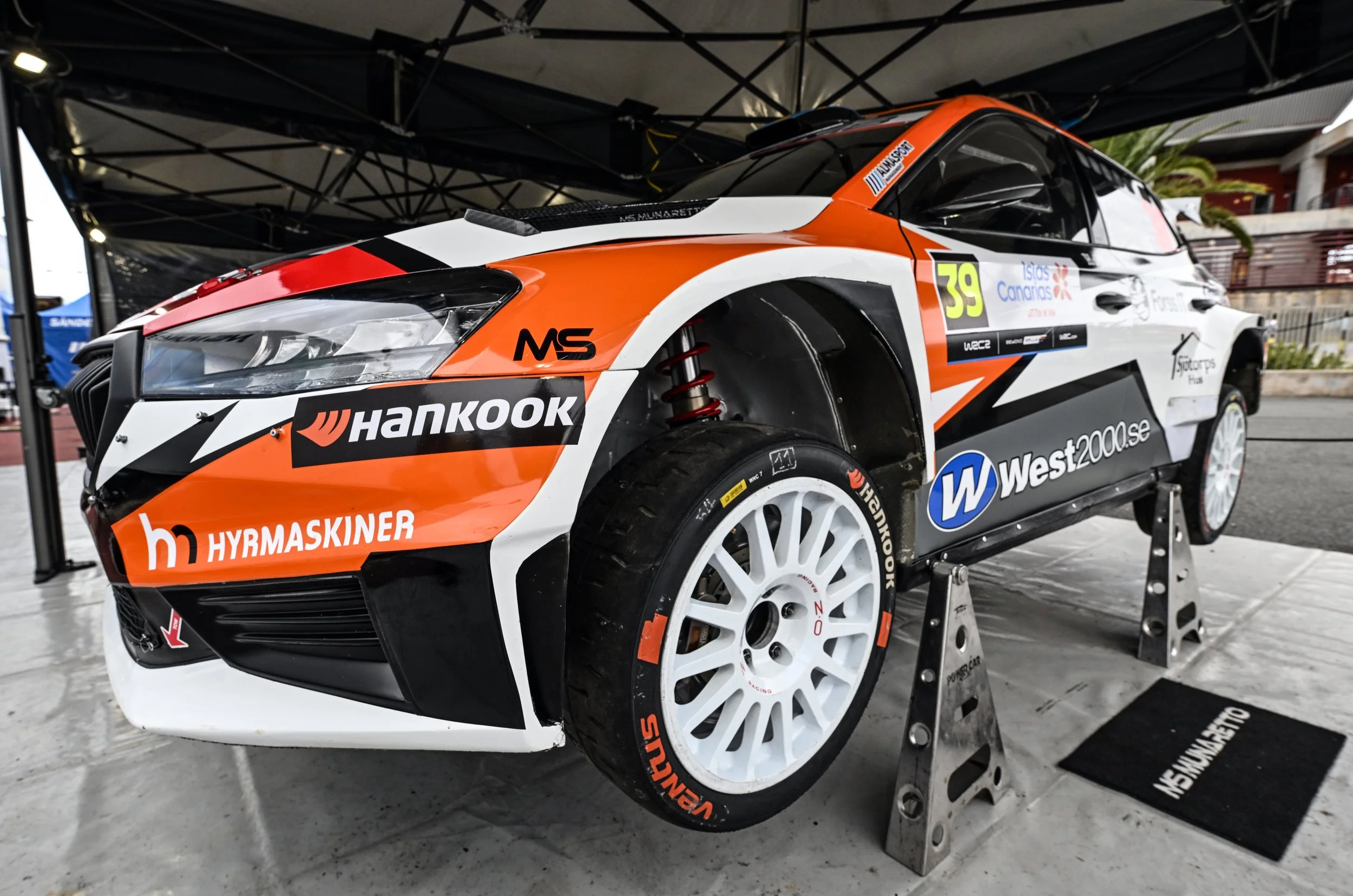 A rally car in racing livery, primarily white with orange and black accents, is parked on a stand under a canopy. The car features logos including Hankook, West200.se, and others, with the number 39 on the side. The wheel is visible along with part o