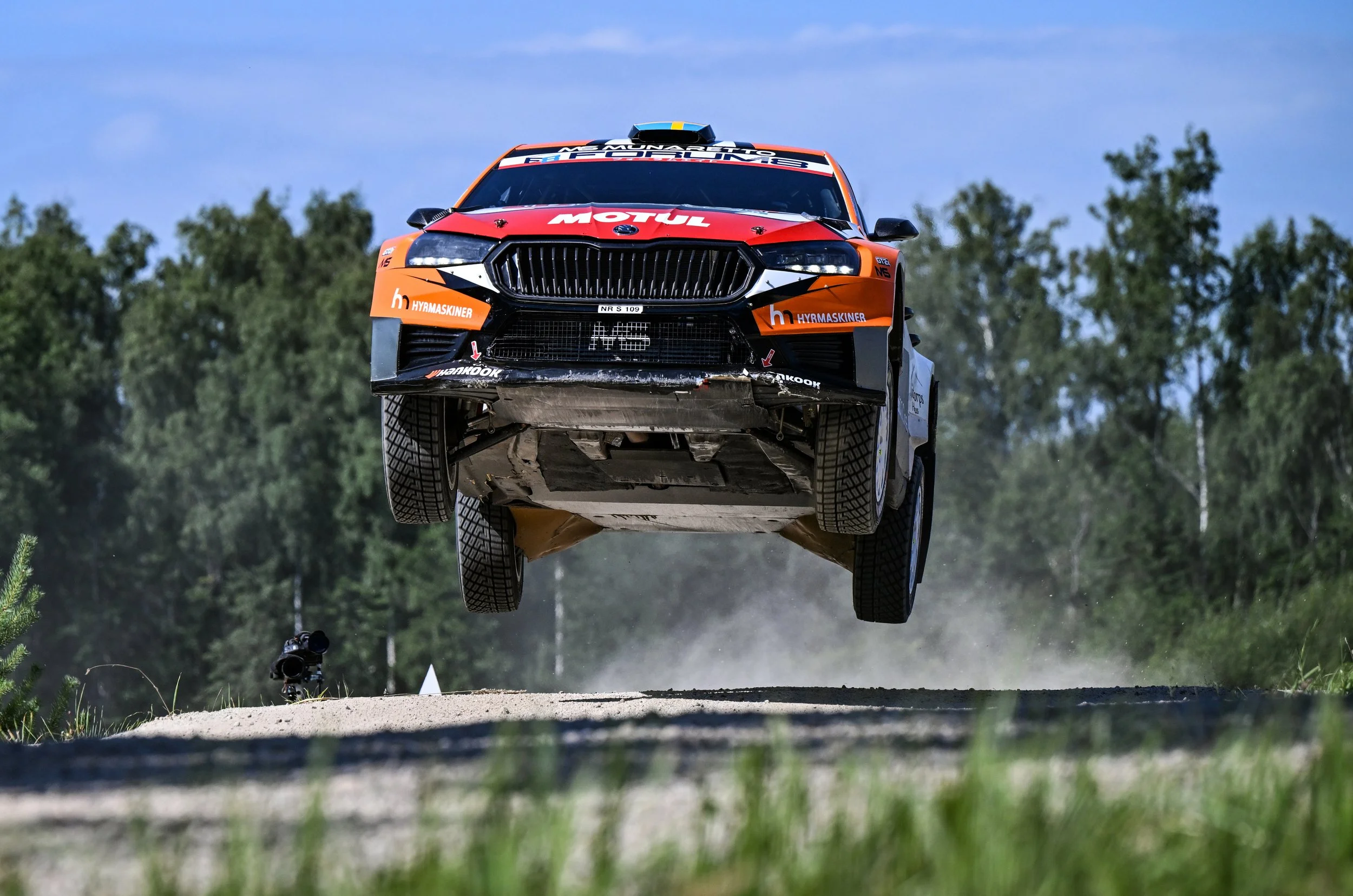 A rally car in orange, black, and white paint flying through the air over a dirt track with trees in the background.