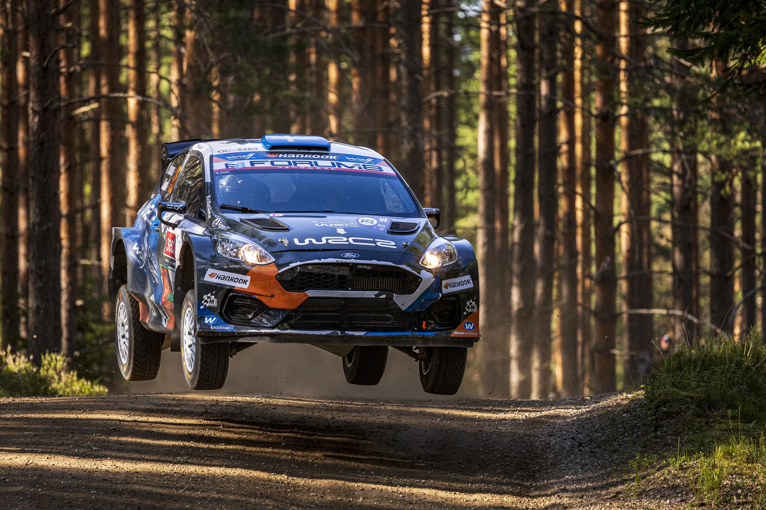 A rally car flying through the air on a dirt road in a forested area during daytime.