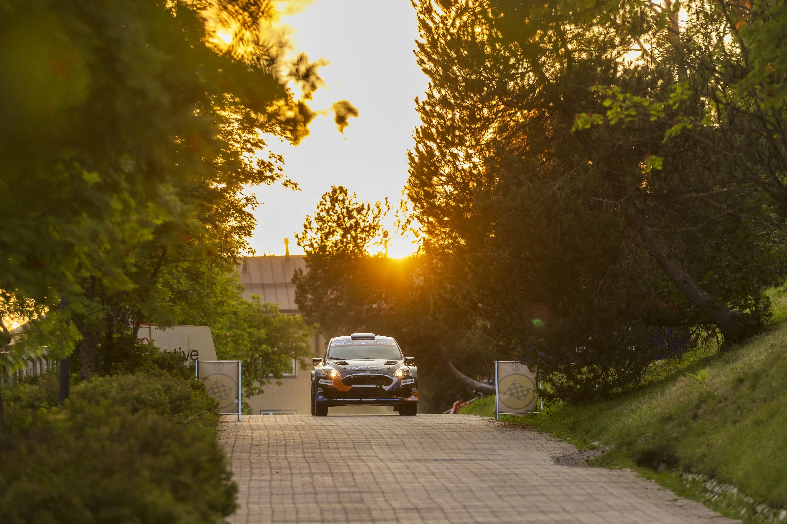 A police car driving on a brick-paved road with trees on either side during sunset, with the sun partially visible behind the trees.