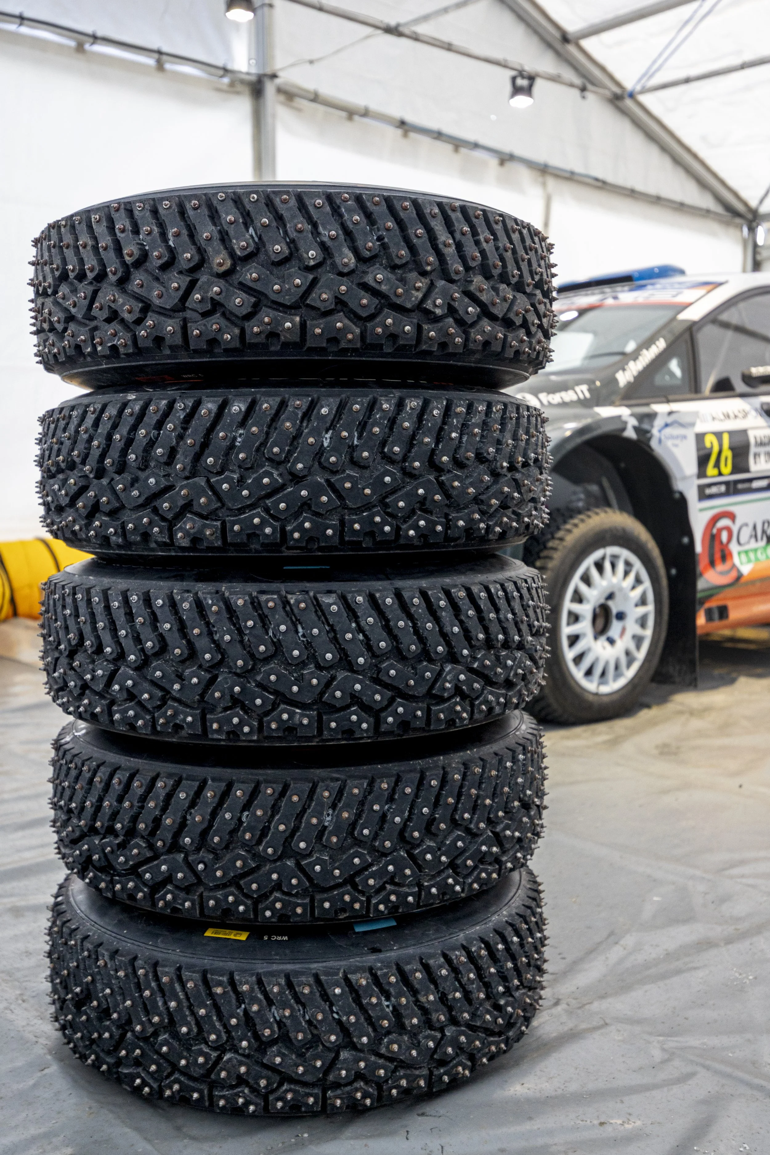 Stack of six racing tires with studded treads in an indoor garage, with a rally car partially visible in the background.