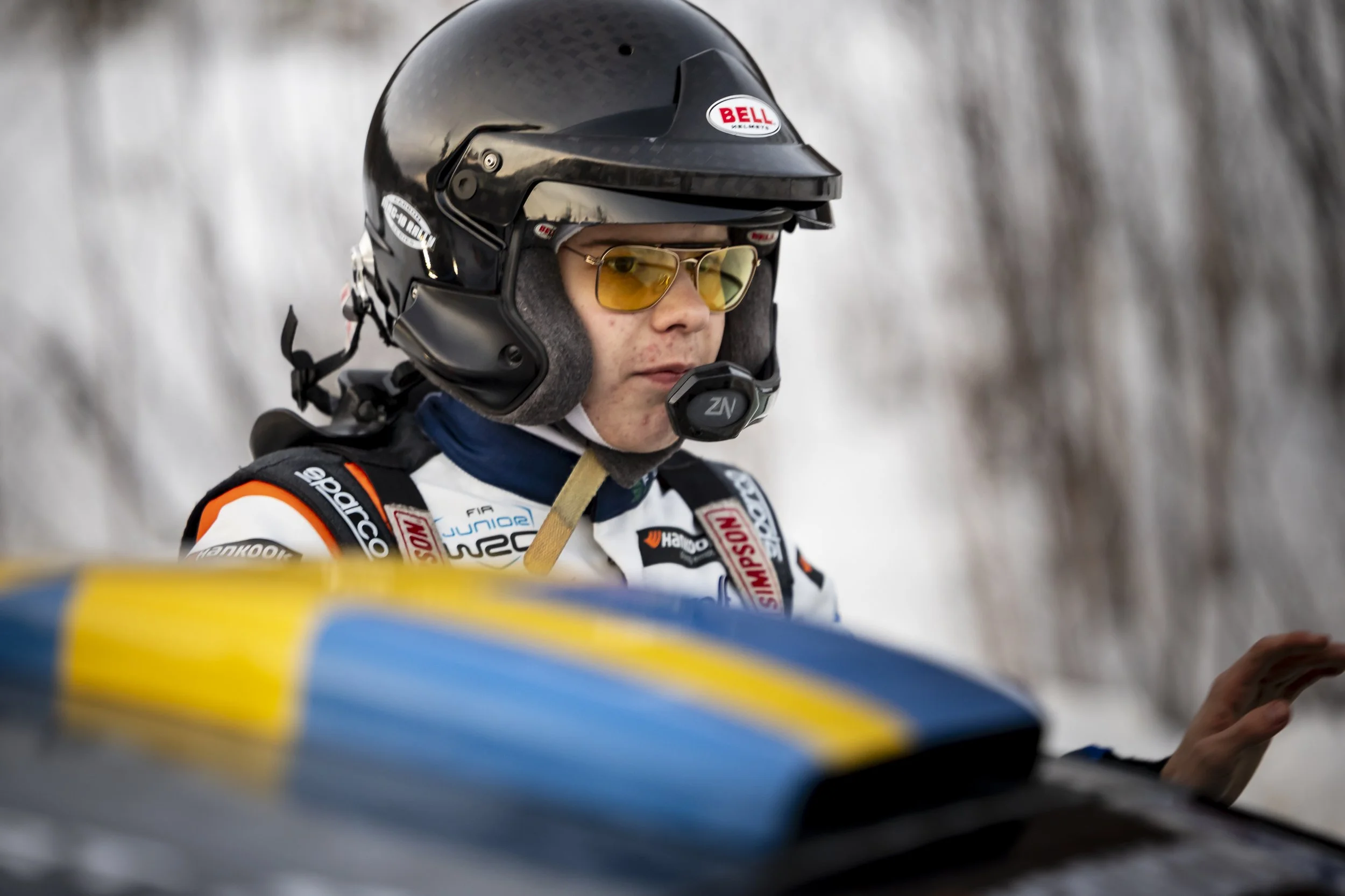 A race car driver wearing a black Bell helmet with yellow tinted sunglasses, dressed in a white racing suit with sponsor patches, sitting in a racing vehicle with a blurred background of trees or natural scenery.
