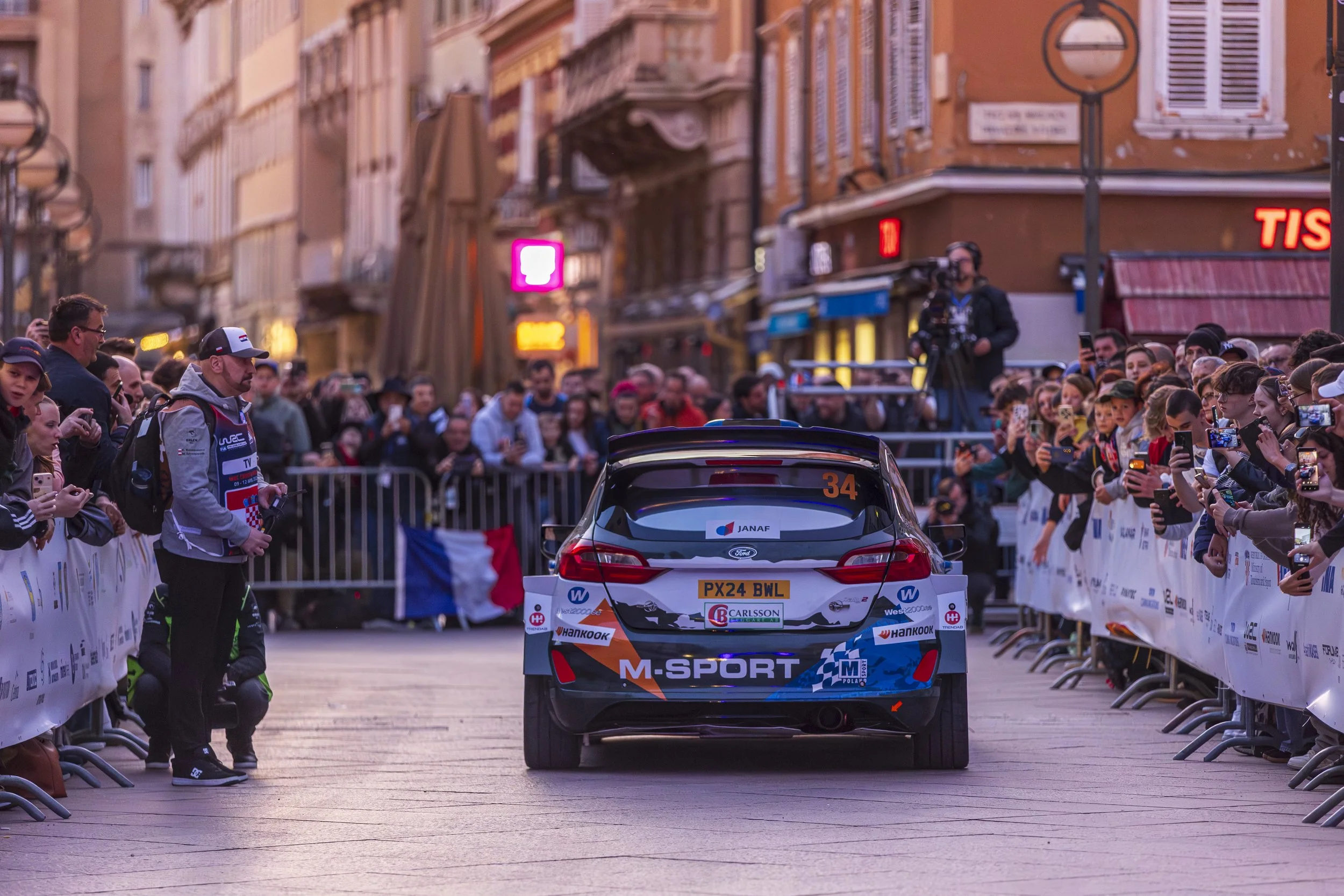 A rally car on a city street with a crowd behind barriers on both sides, photographers and fans taking pictures, buildings with shop signs in the background, and a person with a camera on a platform near the car.