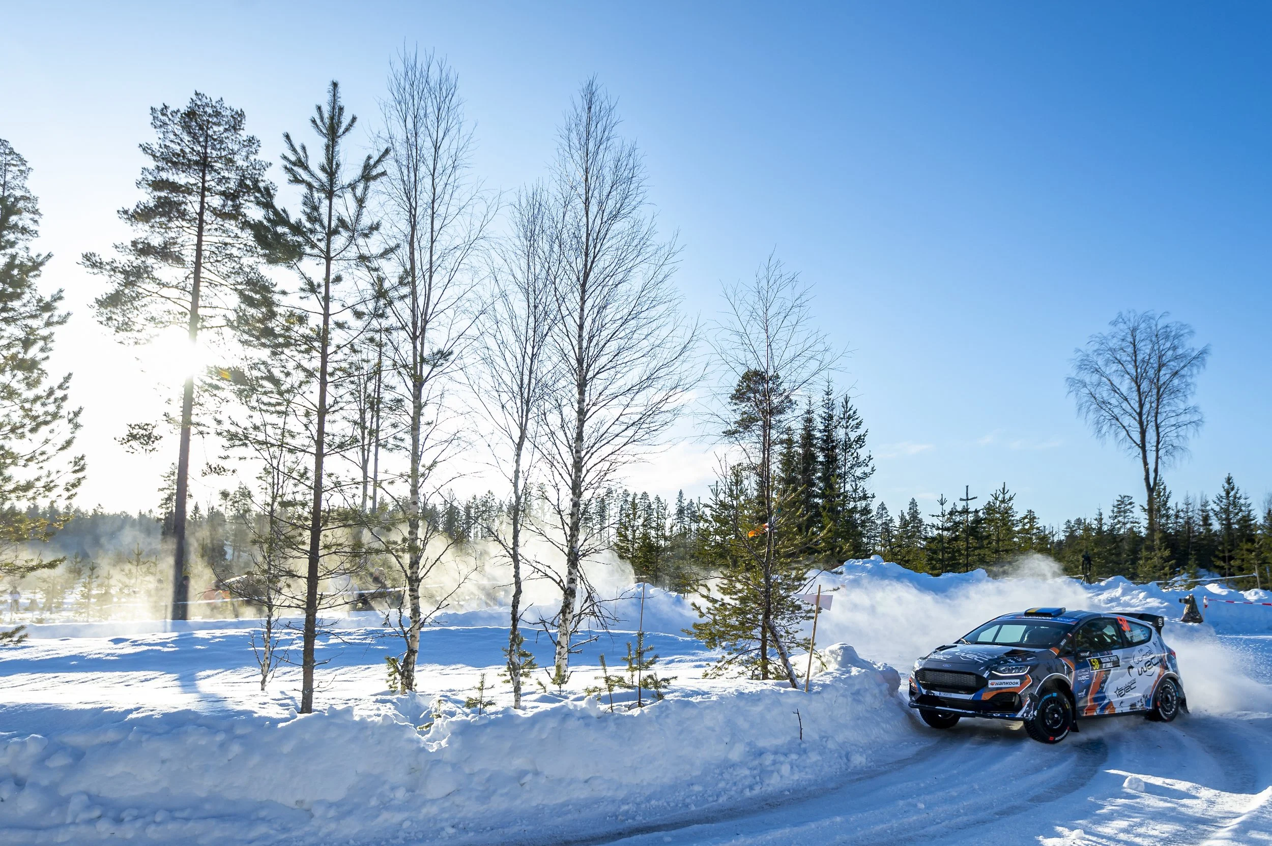 A snowy landscape with a line of tall, leafless trees and evergreen trees against a clear blue sky. In the foreground, there are parked race cars with racing decals, and steam or exhaust rising from the snowy ground.