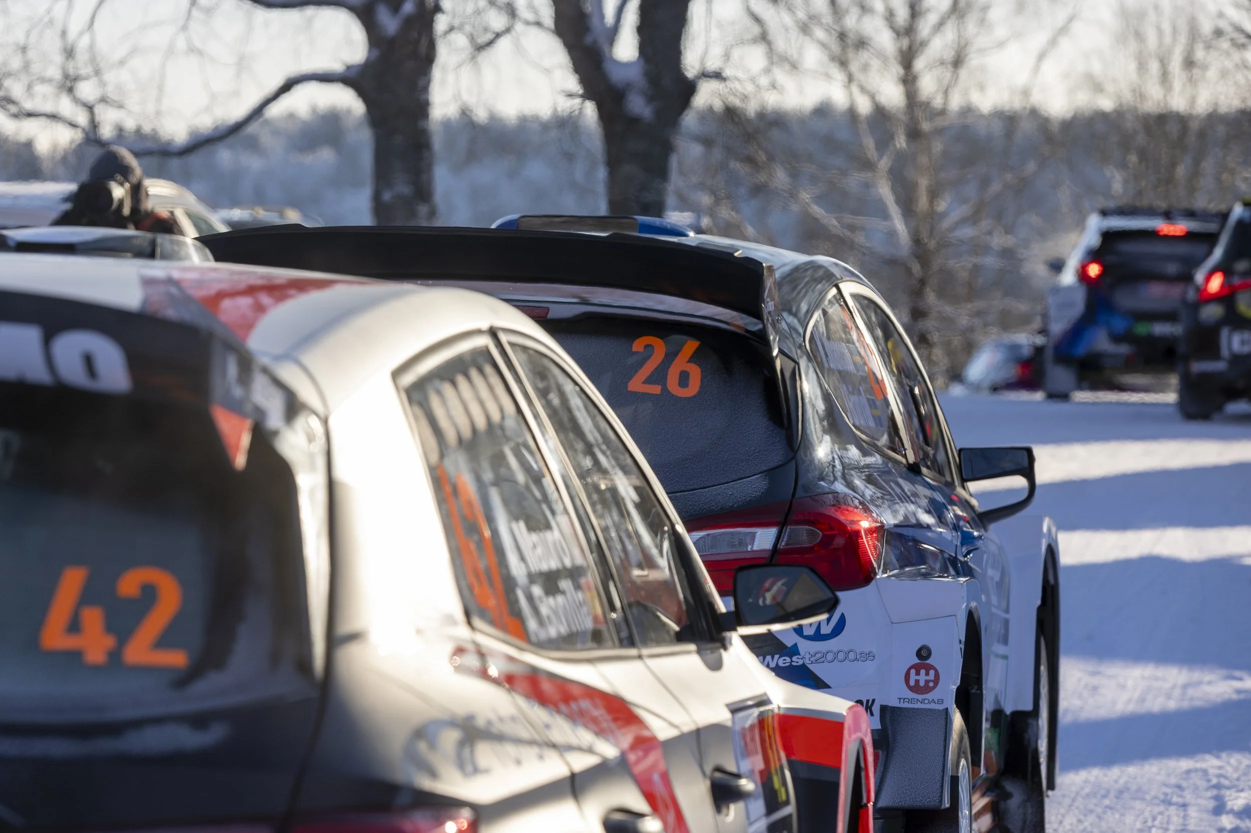 Line of race cars parked on snow-covered ground with leafless trees in the background.